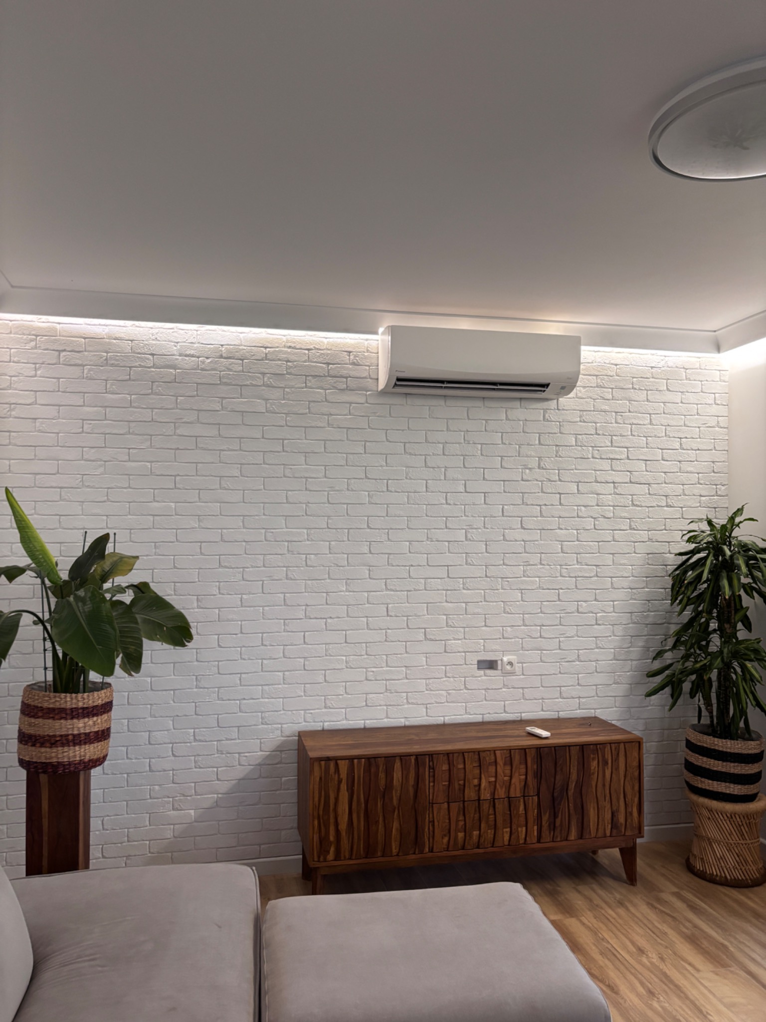 Klimatyzator na białej ceglanej ścianie z oświetleniem LED, drewniana komoda, rośliny doniczkowe i szara sofa w nowoczesnym salonie.