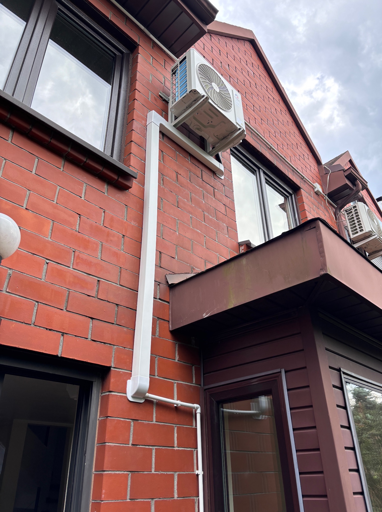 Zewnętrzna jednostka klimatyzacji zamontowana na ceglanej ścianie domu z brązowymi oknami i dachem. Widoczna biała rura odprowadzająca skropliny.