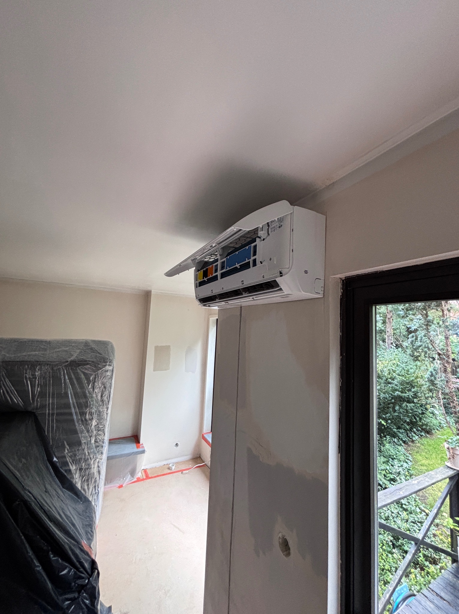 Biały klimatyzator ścienny z otwartą klapą w jasnym wnętrzu w trakcie remontu, widok na zewnątrz przez okno. Meble zabezpieczone folią.