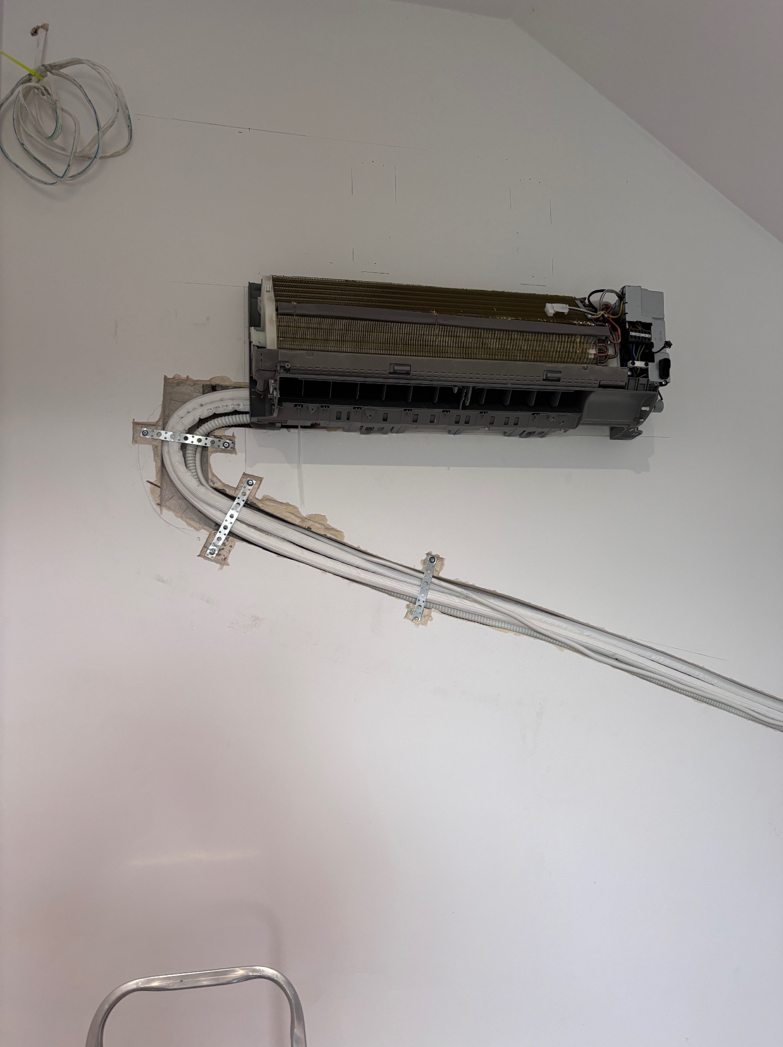 Montaż klimatyzatora ściennego z widocznymi rurami przymocowanymi do ściany za pomocą metalowych uchwytów. Widoczna drabina i przewody.