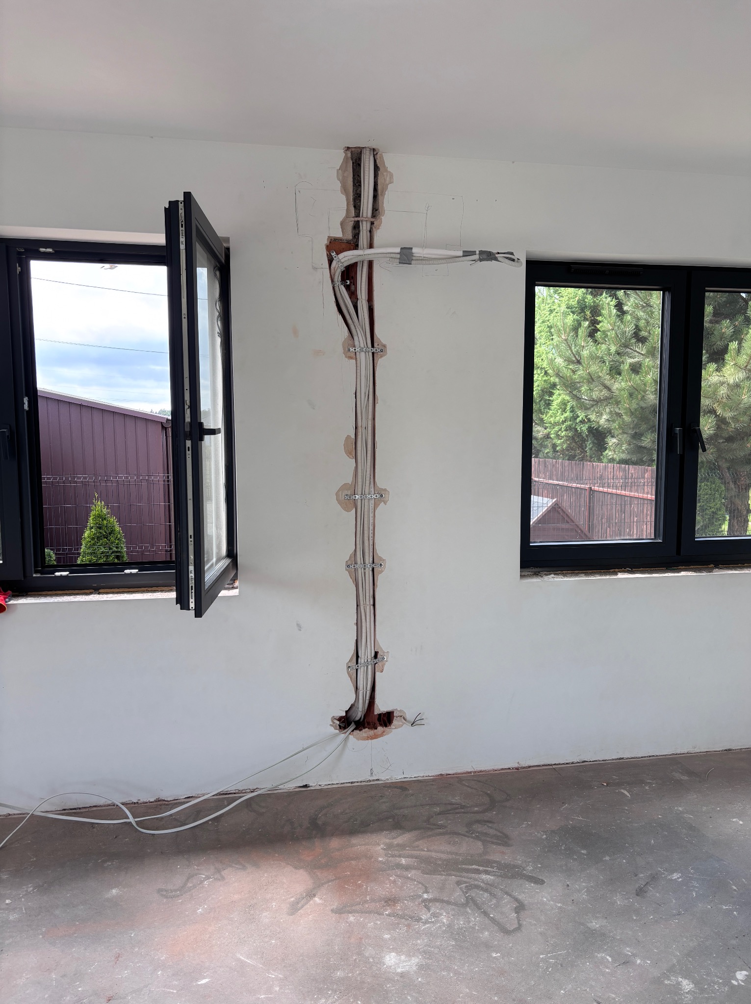 Wewnętrzna instalacja przewodów elektrycznych w ścianie, widoczne okna z czarnymi ramami i fragmenty krajobrazu za nimi. Przewody zamocowane klamrami.