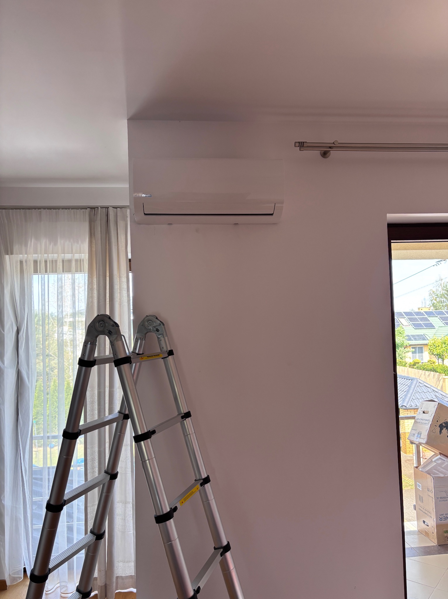 Biały klimatyzator na ścianie w pomieszczeniu, drabina aluminiowa oparta o ścianę, kartony z klimatyzacją przy drzwiach. Widok na dom z panelami słonecznymi.