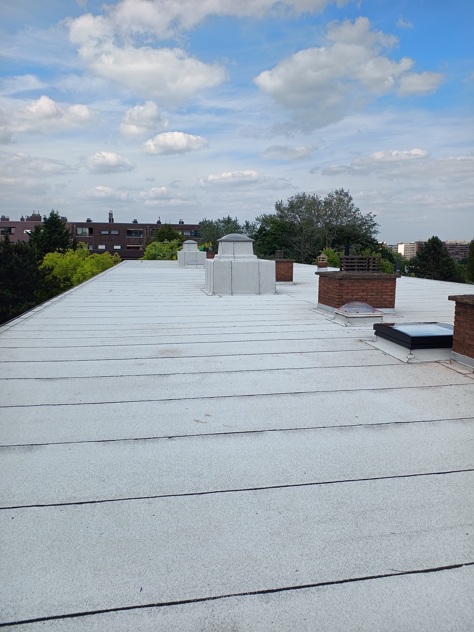 Biały dach płaski z kominami i świetlikami na tle błękitnego nieba z chmurami. Widok z góry na budynki w oddali. Pokrycie dachu z papy.