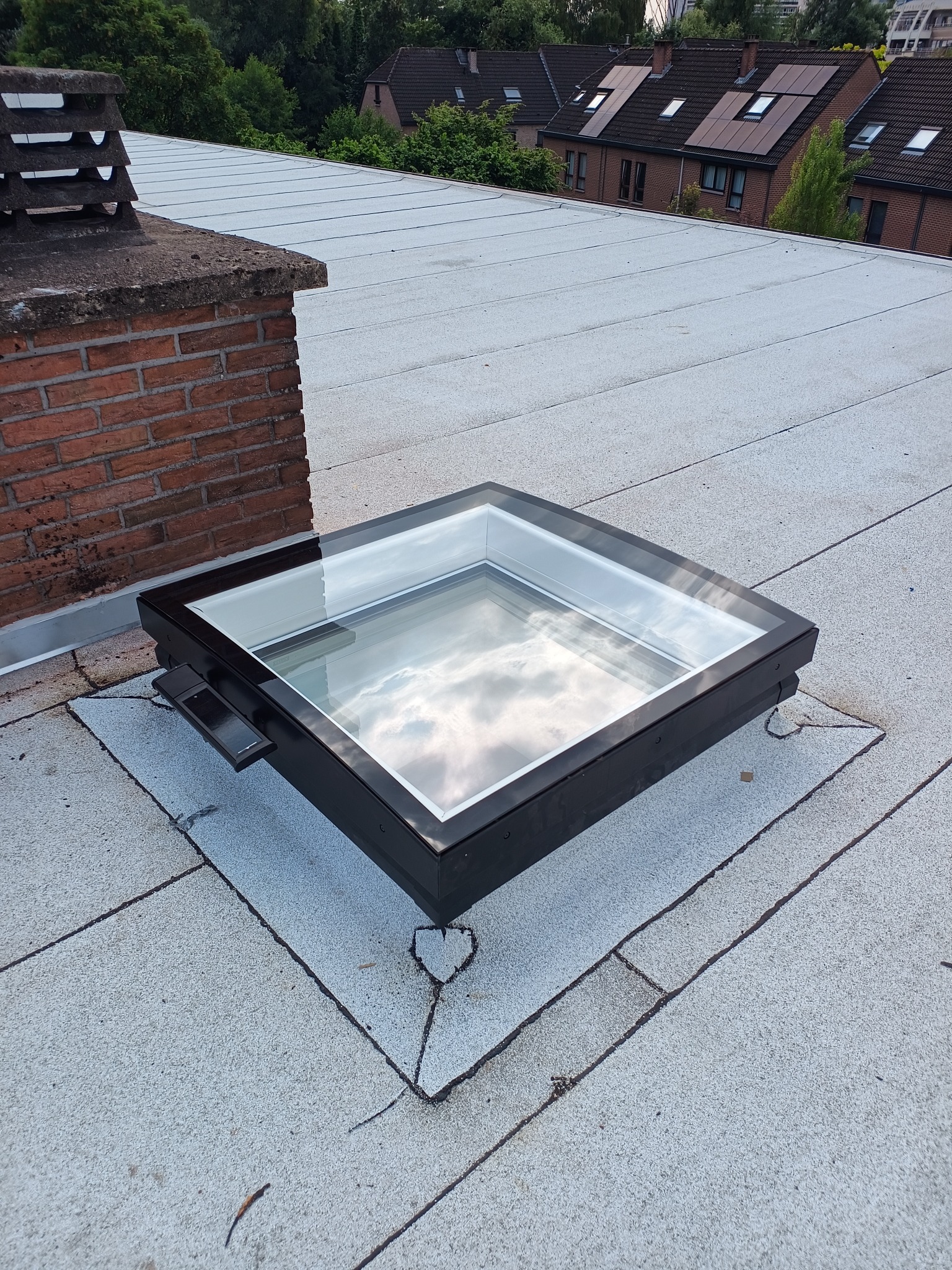 Płaski dach pokryty papą z zamontowanym, czarnym świetlikiem dachowym. W tle komin z cegły i budynki mieszkalne.