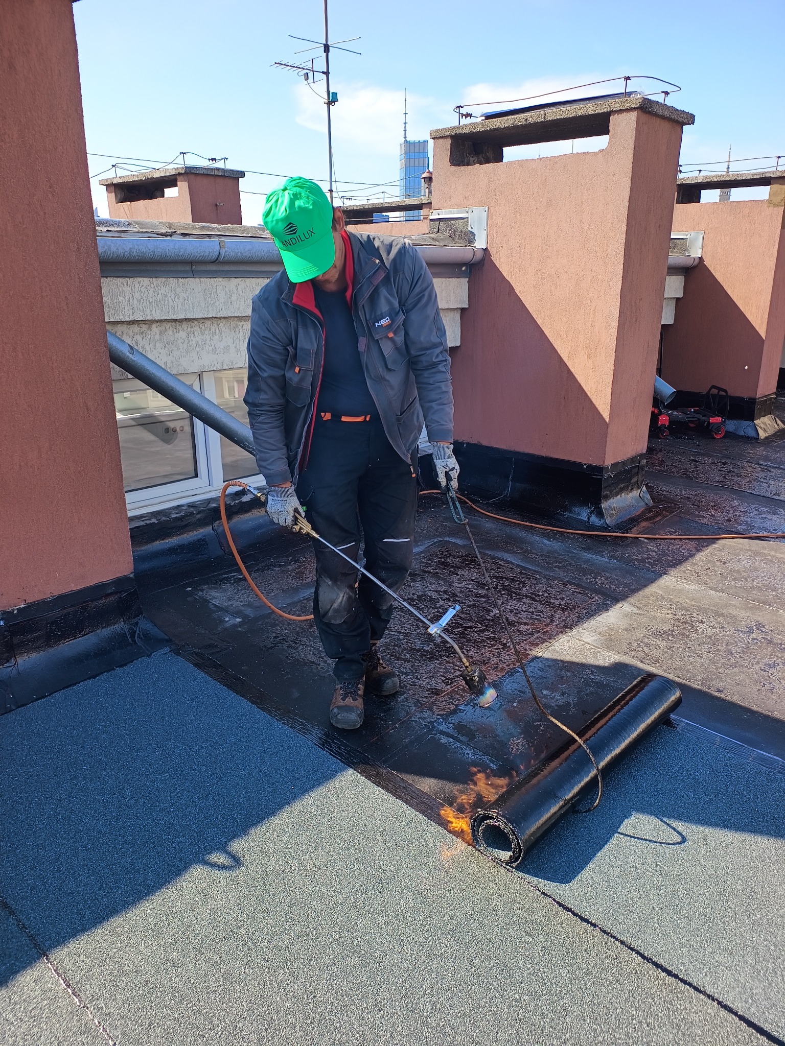 Pracownik w zielonej czapce topi papę na dachu budynku, używając palnika. Widoczne kominy, fragment rynny i panorama miasta w tle. Etap prac dekarskich.