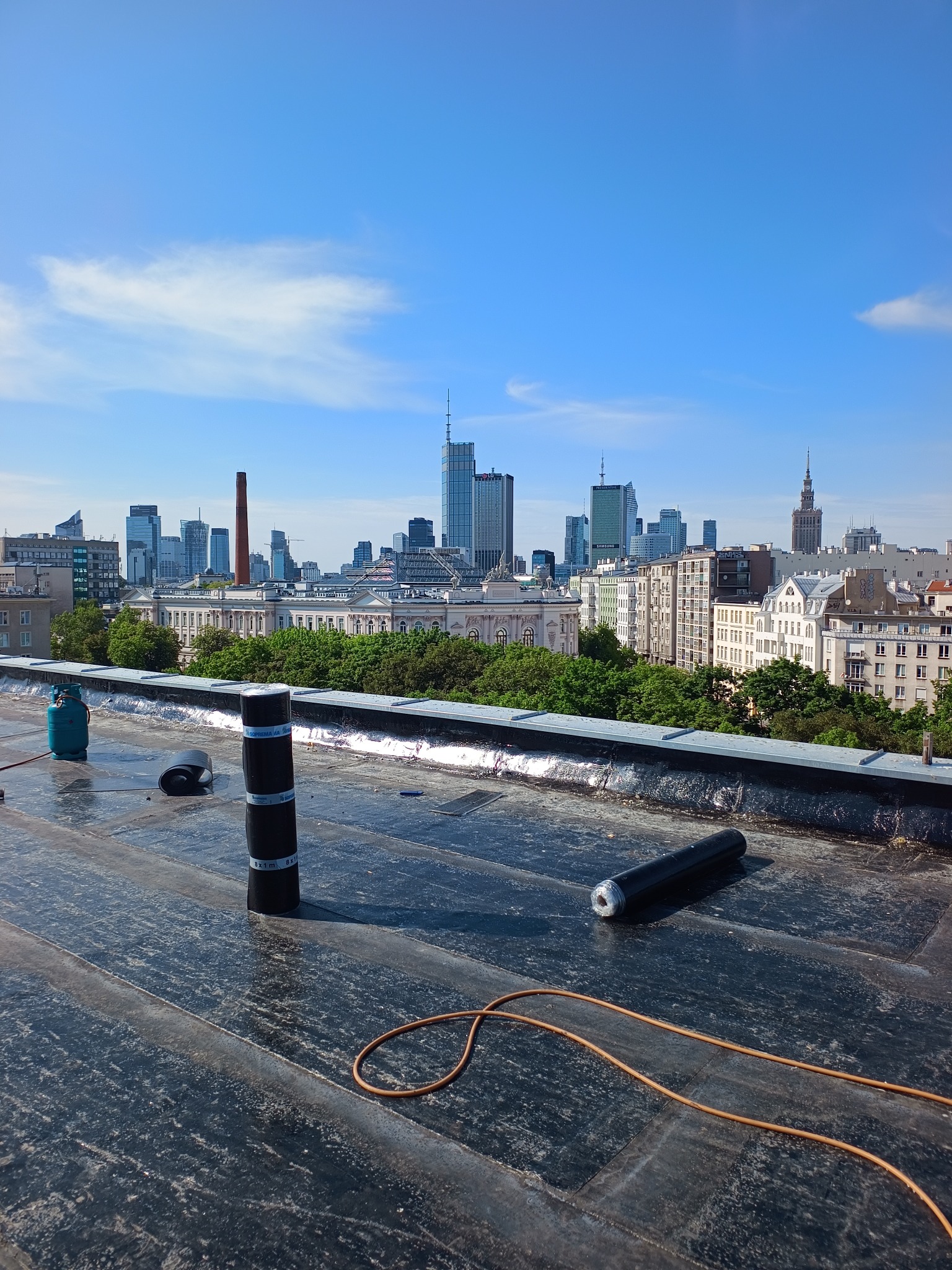 Prace dekarskie na dachu z widokiem na panoramę Warszawy. Rolki papy, butla gazowa i narzędzia dekarskie na tle miejskiego pejzażu w słoneczny dzień.