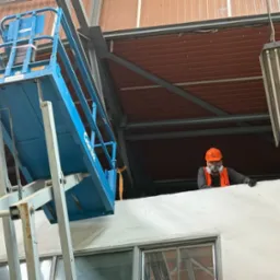 Pracownik w masce, na podnośniku Genie, maluje ścianę wewnątrz budynku. Widoczny sufit z blachy trapezowej i oświetlenie przemysłowe.