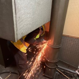 LUGINVEST SPÓŁKA Z OGRANICZONĄ ODPOWIEDZIALNOŚCIĄ - Pracownik w kamizelce tnący metalową rurę szlifierką kątową, iskry rozświetlają ciemne pomieszczenie. Widoczny fragment wentylacji i perforowana ściana.