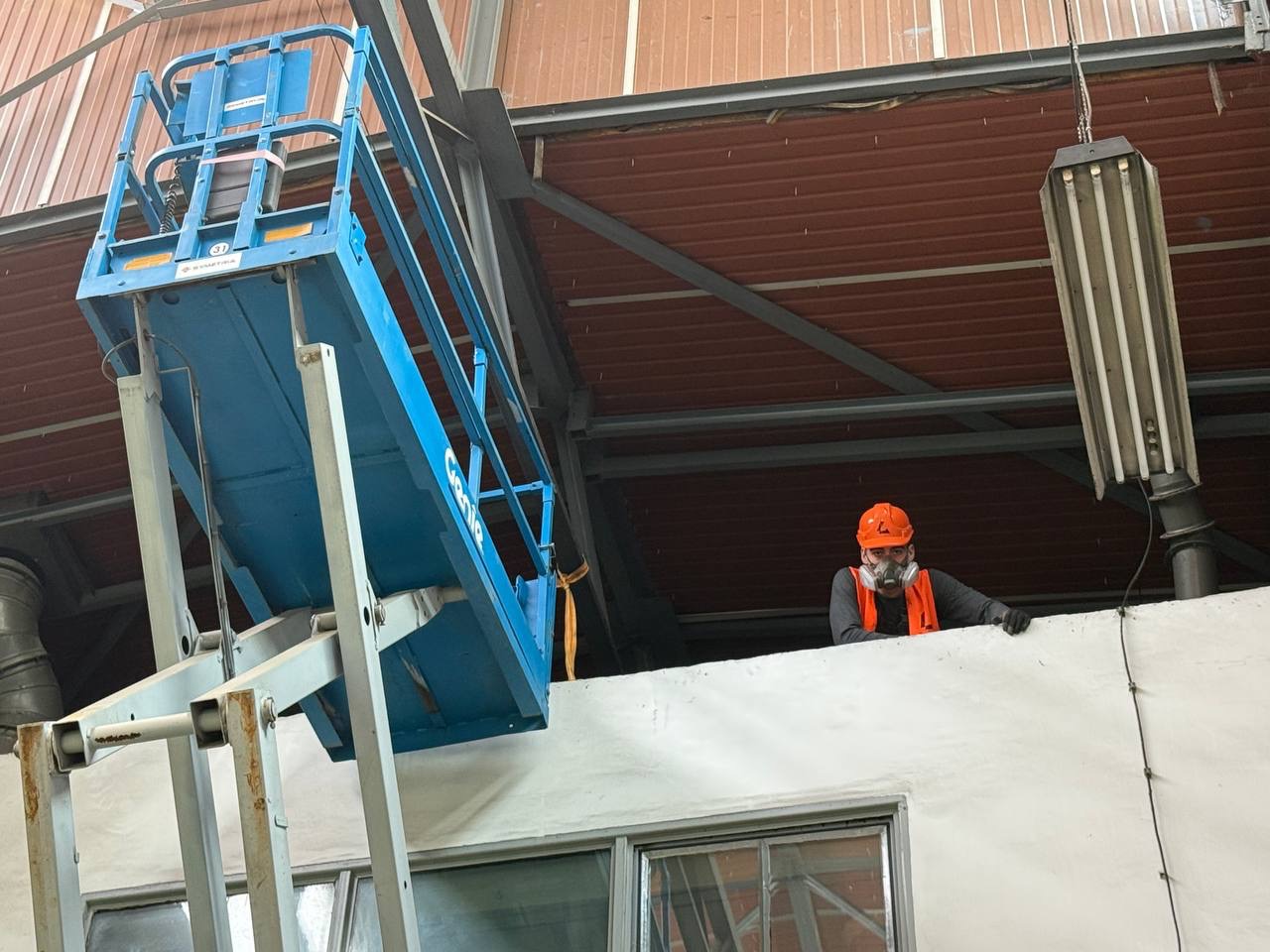 Pracownik w masce, na podnośniku Genie, maluje ścianę wewnątrz budynku. Widoczny sufit z blachy trapezowej i oświetlenie przemysłowe.