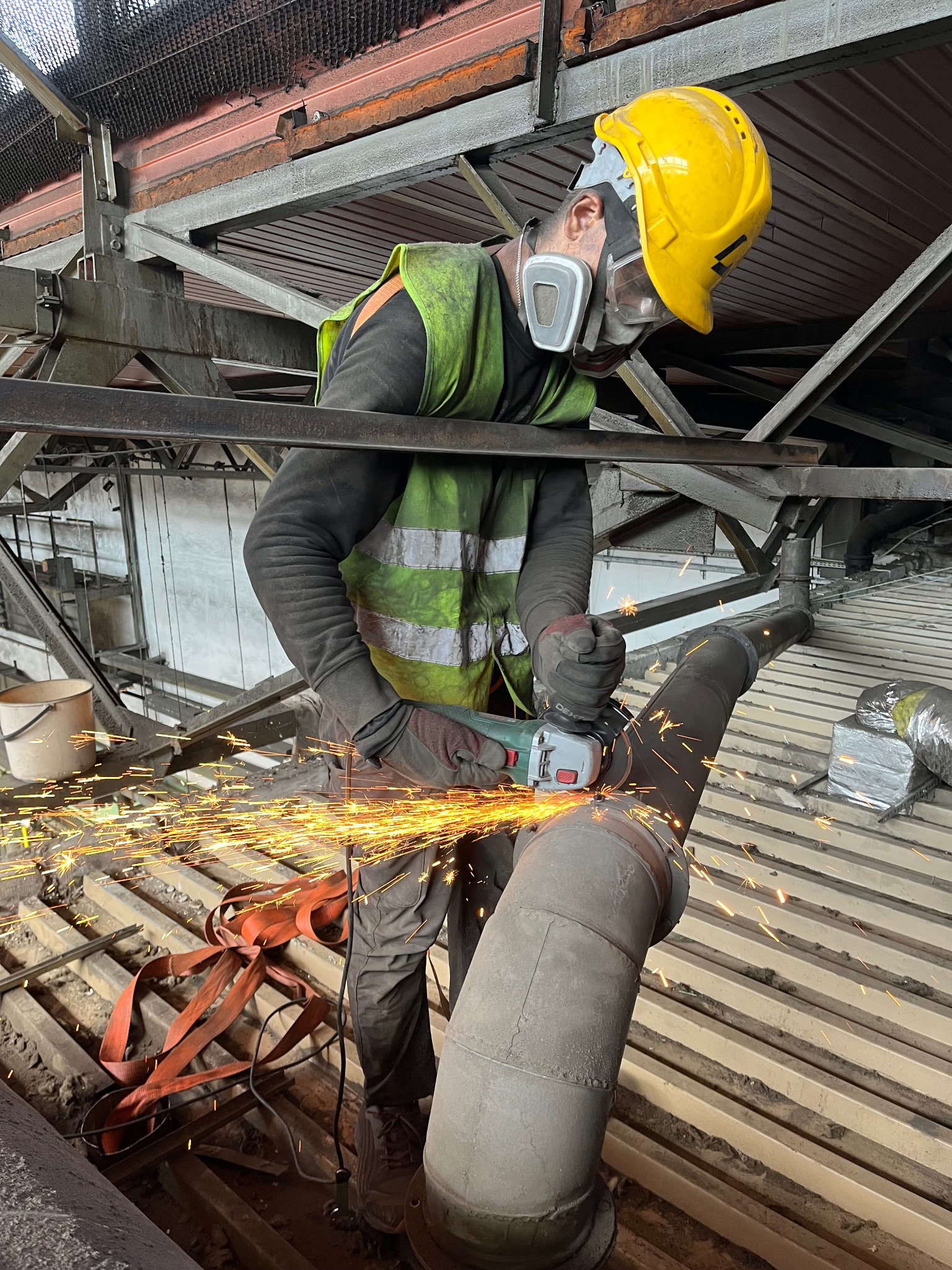 Pracownik w kasku i masce tnący rurę szlifierką kątową, iskry w powietrzu, widoczne elementy konstrukcyjne budynku i podłoga z metalowych żeber.
