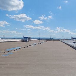 Semplex Service Group Sp. z.o.o. - Szeroki, płaski dach hali przemysłowej z świetlikami dachowymi i kałużami po deszczu, na tle błękitnego nieba z chmurami. Widoczne elementy infrastruktury.