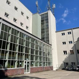 Semplex Service Group Sp. z.o.o. - Budynek biurowy z dużą szklaną fasadą i antenami na dachu, widziany z poziomu dziedzińca w słoneczny dzień. Widoczne rośliny wewnątrz i zaparkowany samochód.