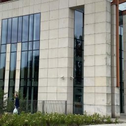 Semplex Service Group Sp. z.o.o. - Elewacja biurowca z dużymi oknami, widoczne ślady zabrudzeń i graffiti na dolnych płytach. Osoba przechodzi obok budynku. Dwie kamery monitoringu na ścianie.