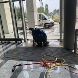 Semplex Service Group Sp. z.o.o. - Automat myjący i akcesoria na mokrej podłodze w holu biurowca, widoczny biały bus za szklaną ścianą. Sprzątanie po umyciu podłogi.