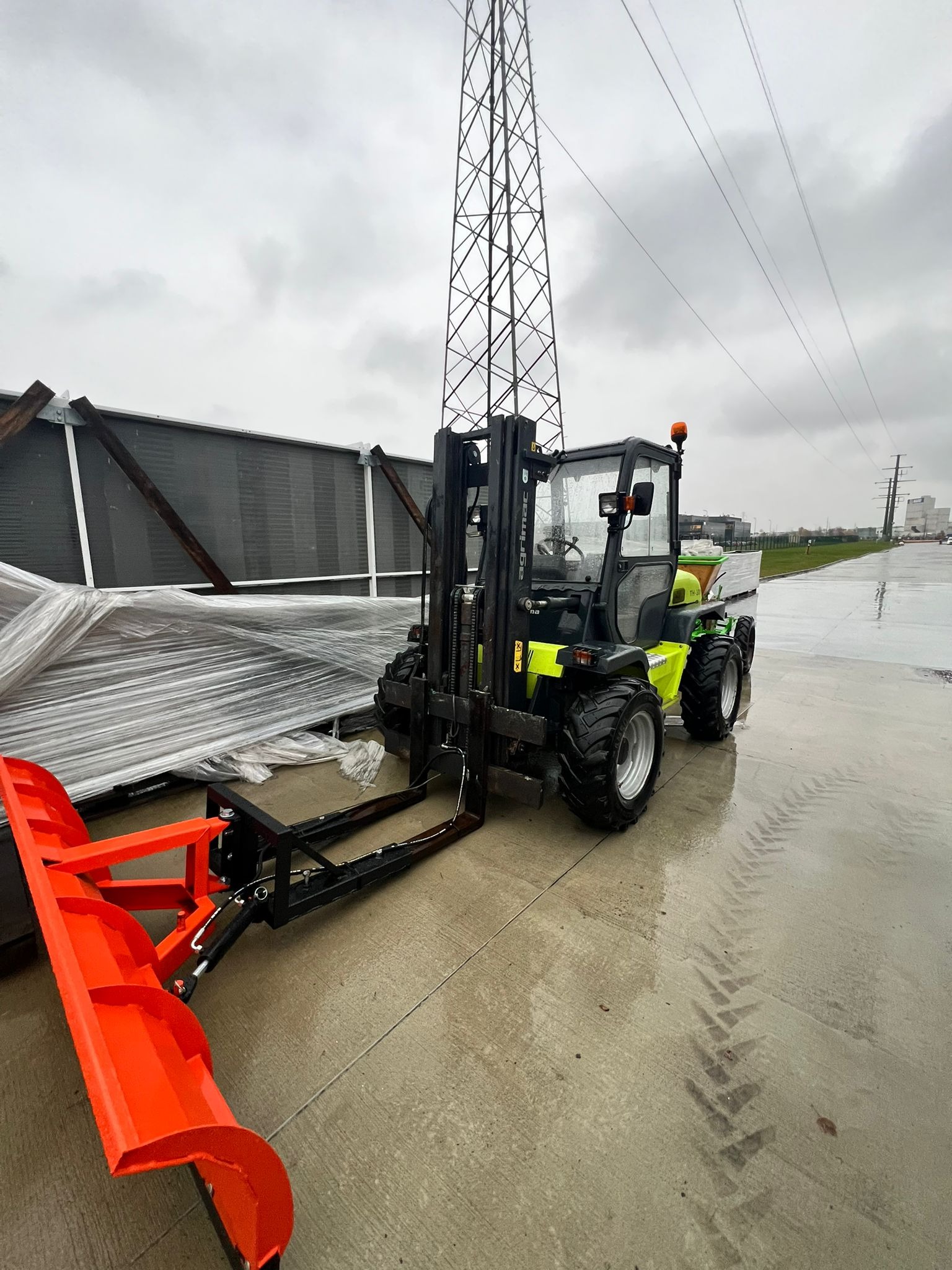 Żółto-czarny wózek widłowy z pomarańczowym pługiem do śniegu na mokrym placu, w tle słup wysokiego napięcia i budynek. Widoczne ślady opon.