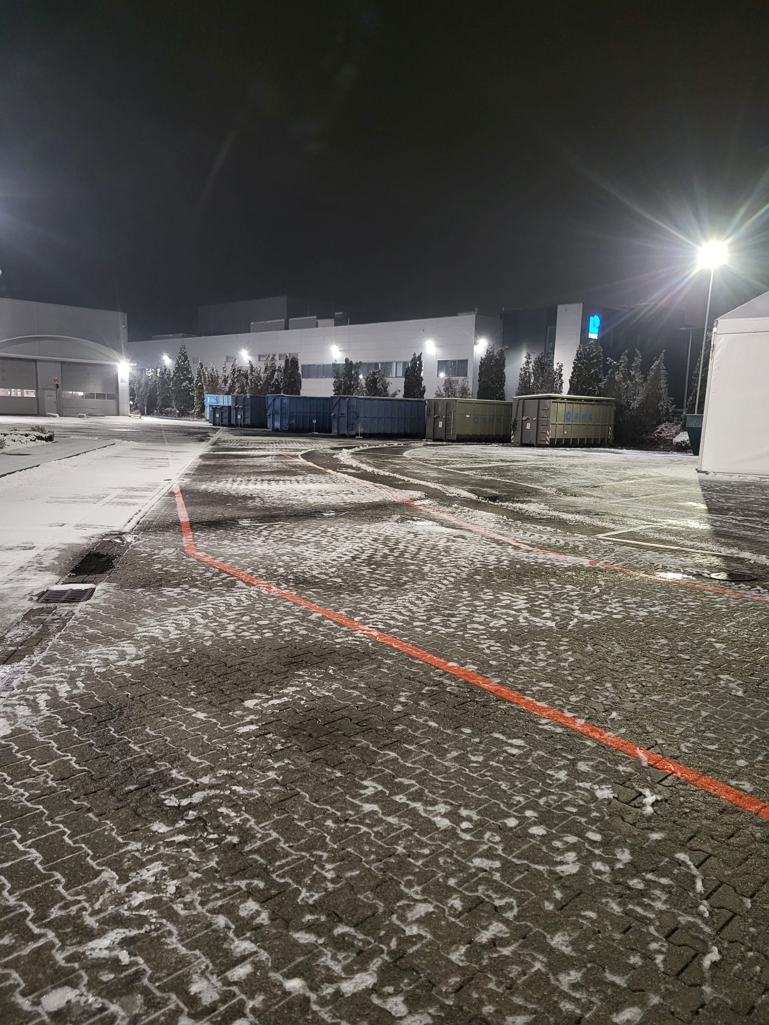 Nocny widok na plac z kostki brukowej, częściowo pokryty śniegiem. Widoczne kontenery, budynek w tle i oświetlenie uliczne. Czerwone linie wyznaczają pasy.