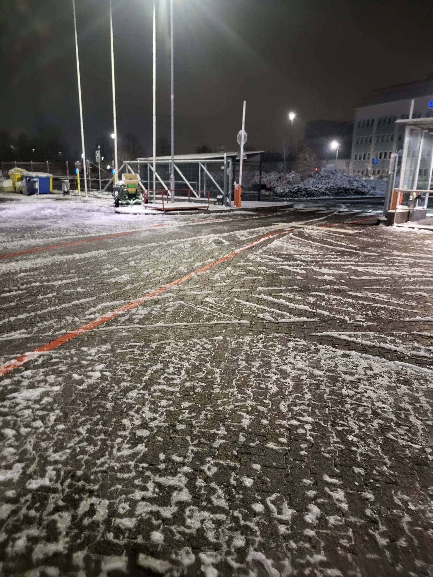 Nocne odśnieżanie kostki brukowej na parkingu z użyciem traktorka. Widoczne ślady po odśnieżaniu i resztki śniegu. W tle budynek i oświetlenie.