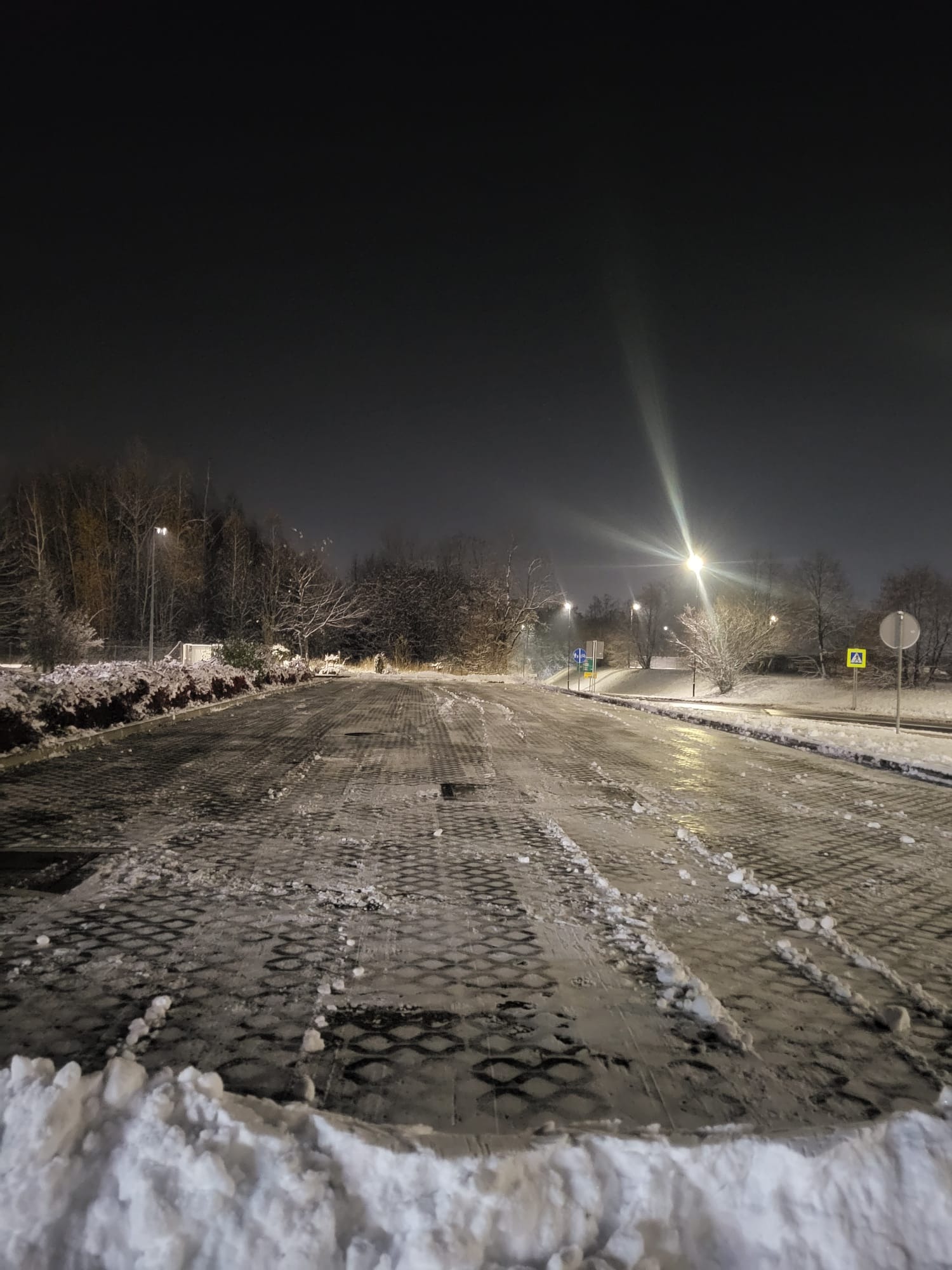 Nocny widok na częściowo odśnieżony parking z kostki brukowej, oświetlony latarniami, z resztkami śniegu po bokach i na kostce. Widoczne znaki drogowe.