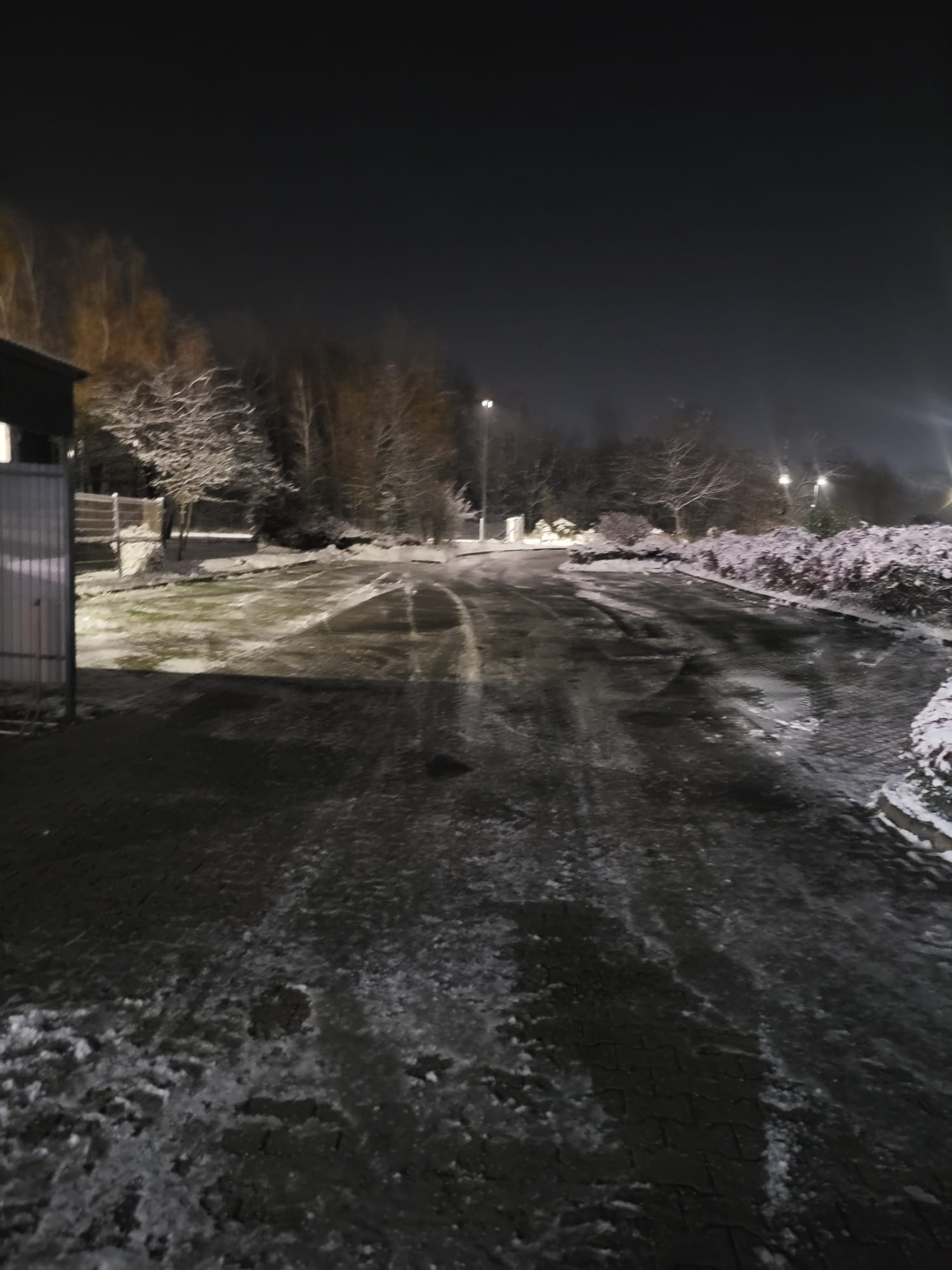 Nocne odśnieżanie parkingu z widocznymi śladami opon, oświetlonego latarniami, z zaspami śniegu przy krawężnikach i drzewach w tle.