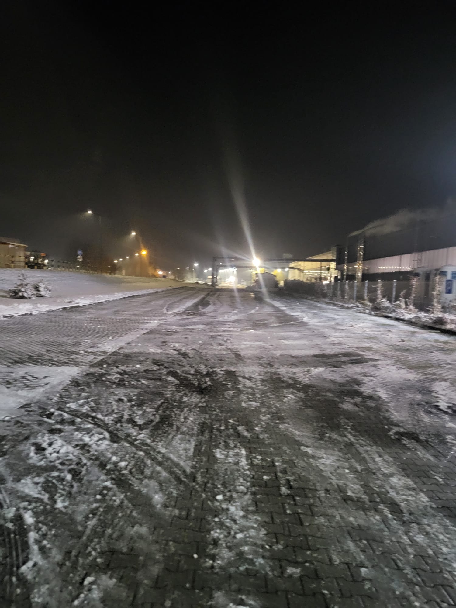 Nocny widok na zasypany śniegiem plac z widocznymi śladami opon, oświetlony latarniami i światłami budynku w tle. Śnieg częściowo stopniał, odsłaniając kostkę brukową.