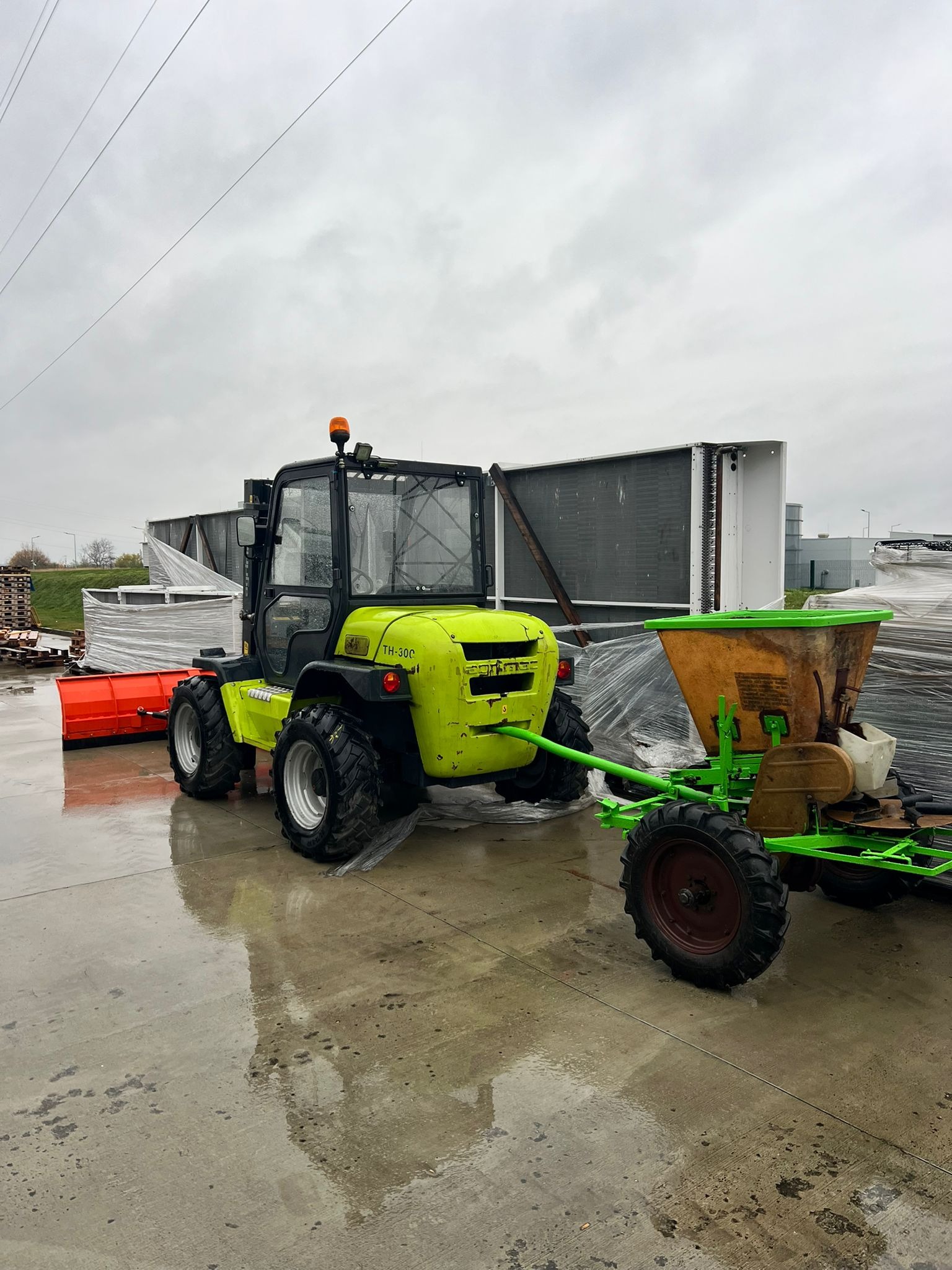 Żółto-czarny traktor TH-300 z pługiem i rozsiewaczem na mokrym placu, w tle palety i klimatyzatory pod folią, pochmurne niebo. Widok z boku, lekko z góry.