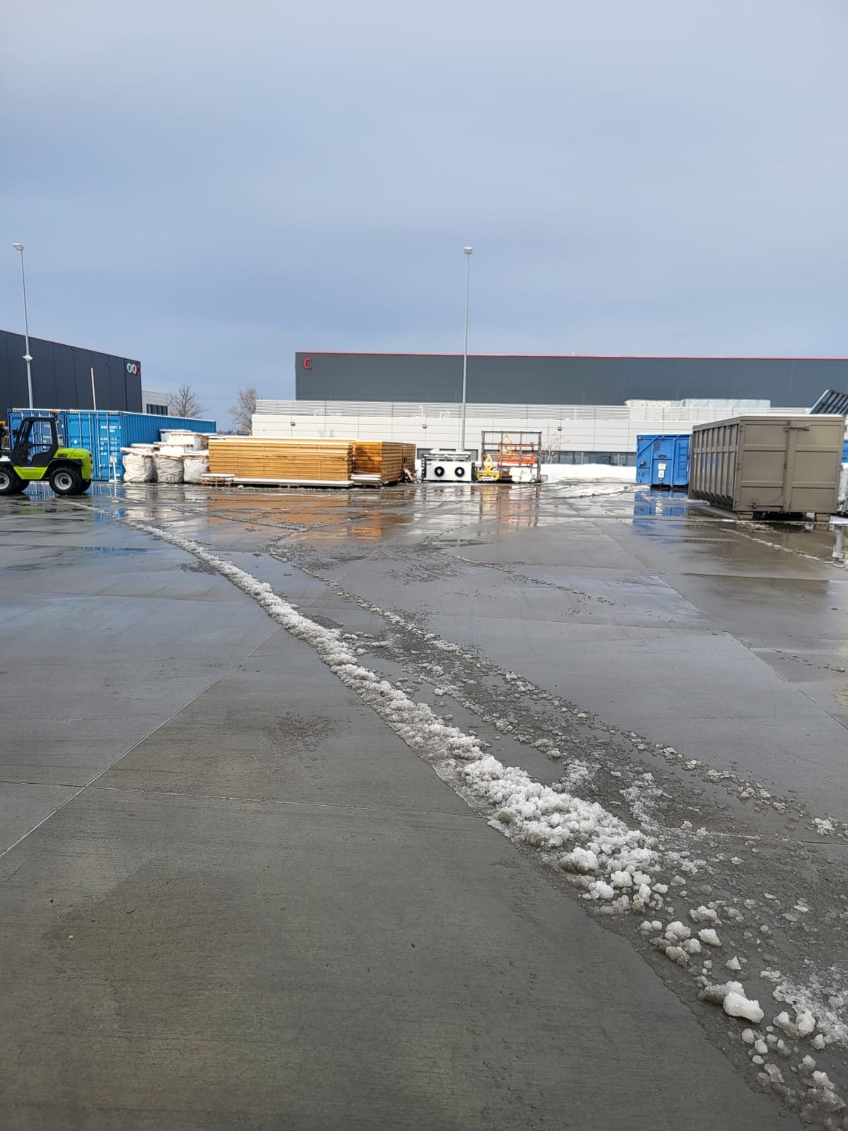Odśnieżony plac magazynowy z widocznymi śladami opon i resztkami śniegu. W tle budynek magazynowy, kontenery i wózek widłowy.