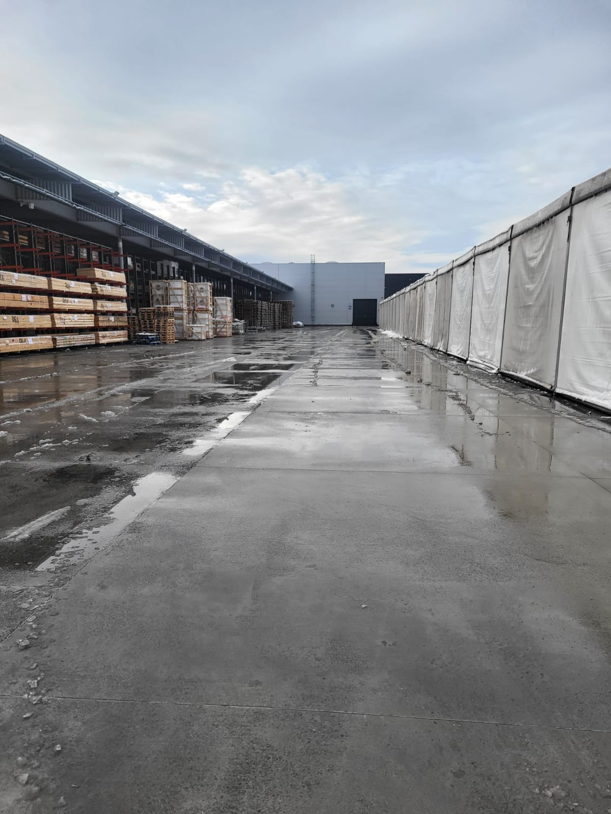 Mokry, betonowy plac magazynowy po roztopach, z widocznymi kałużami i resztkami śniegu. W tle regały z drewnem i paletami, oraz biała hala magazynowa.