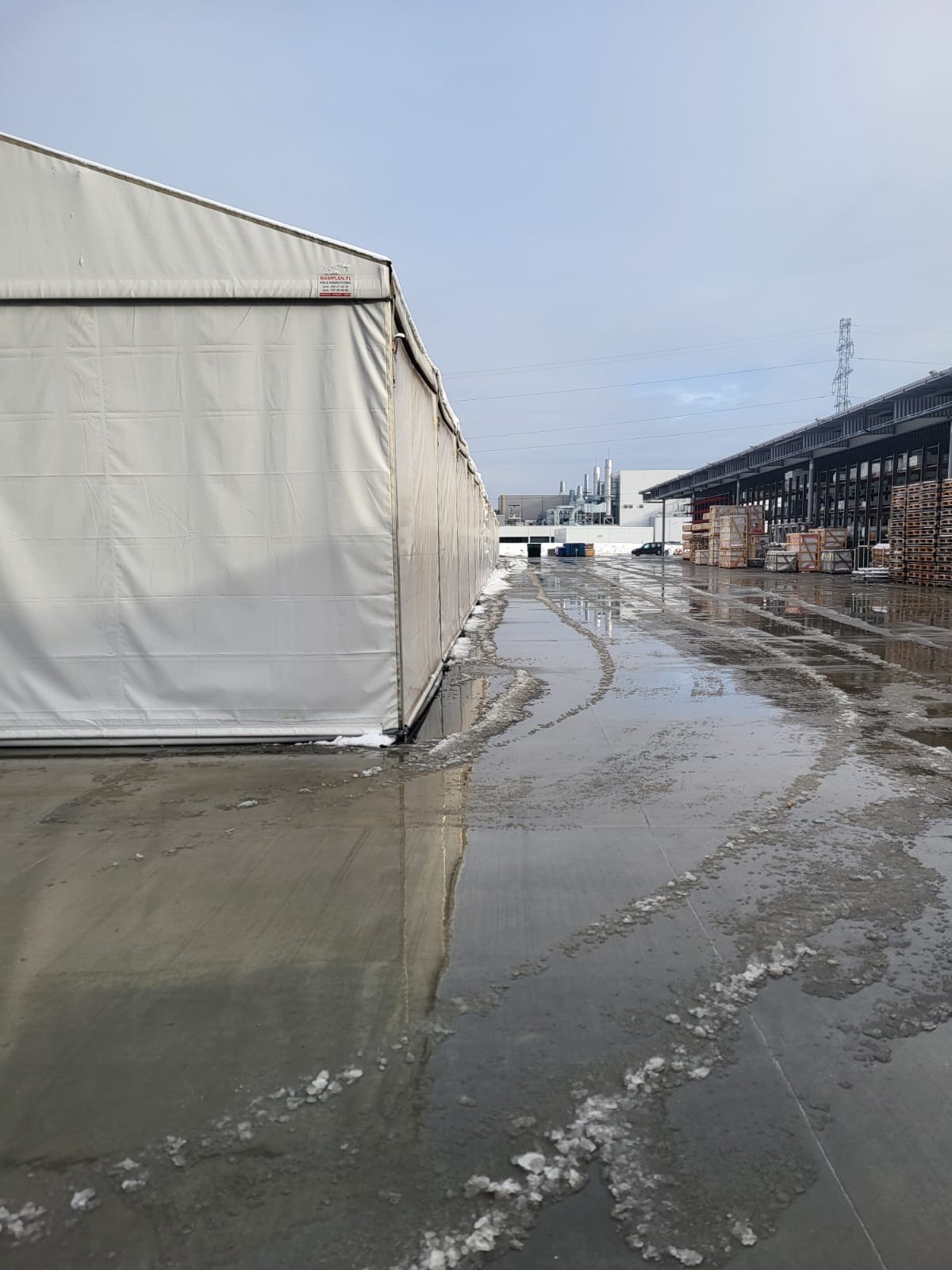 Odśnieżony, mokry plac magazynowy z resztkami śniegu i lodu, widoczny magazyn i hala w tle. Ślady po odśnieżaniu. Perspektywa z dołu.