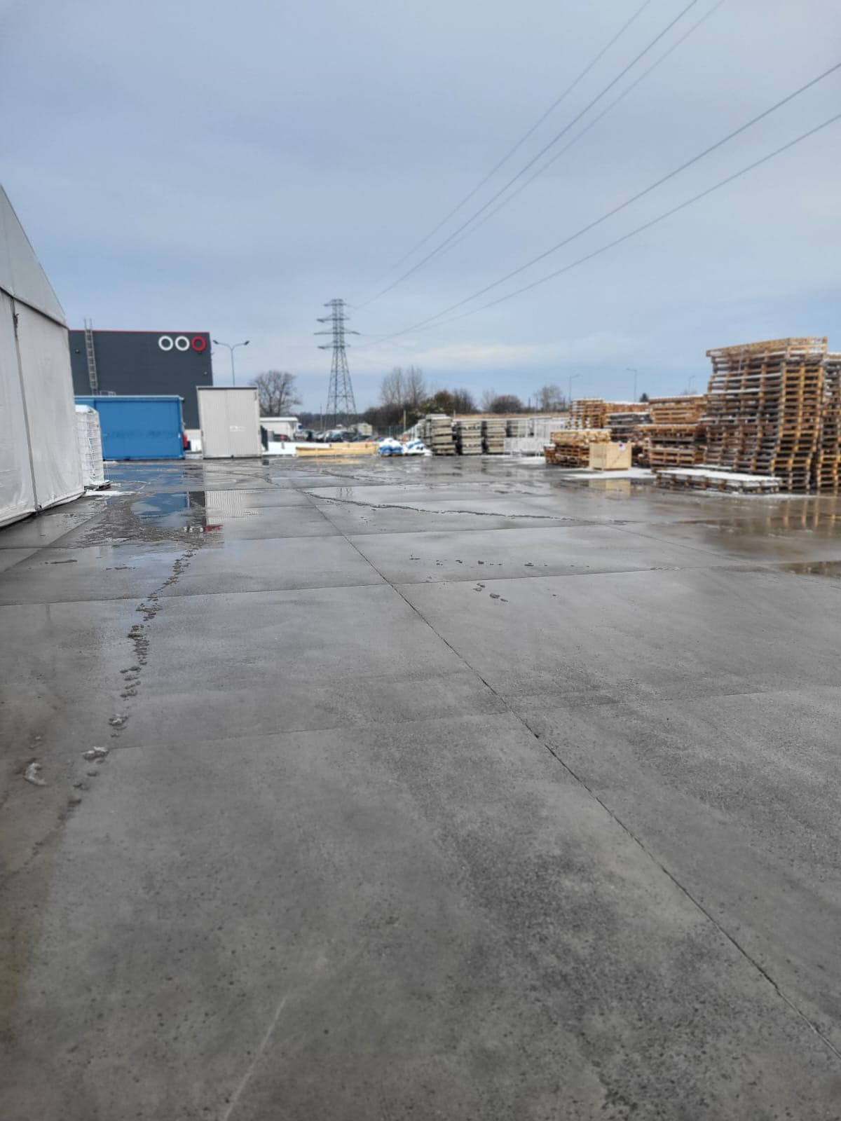 Betonowy plac po odśnieżaniu z paletami w tle, widoczne ślady topniejącego śniegu i kałuże. W oddali budynek z logo i słup wysokiego napięcia.