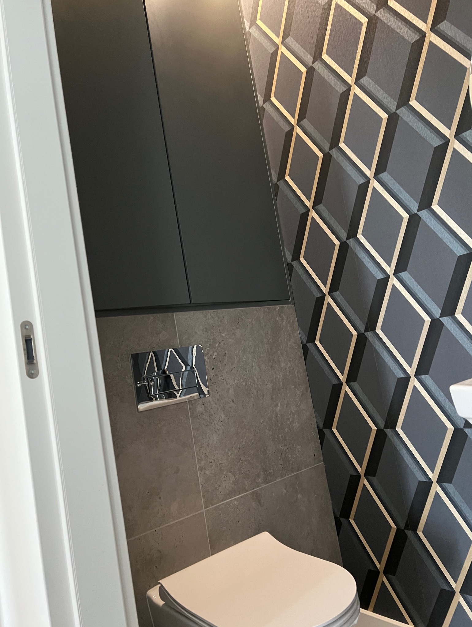 Nowoczesna toaleta po remoncie: grafitowe szafki, geometryczna ściana z akcentami złota i minimalistyczny przycisk spłuczki. Detal wykończenia łazienki.