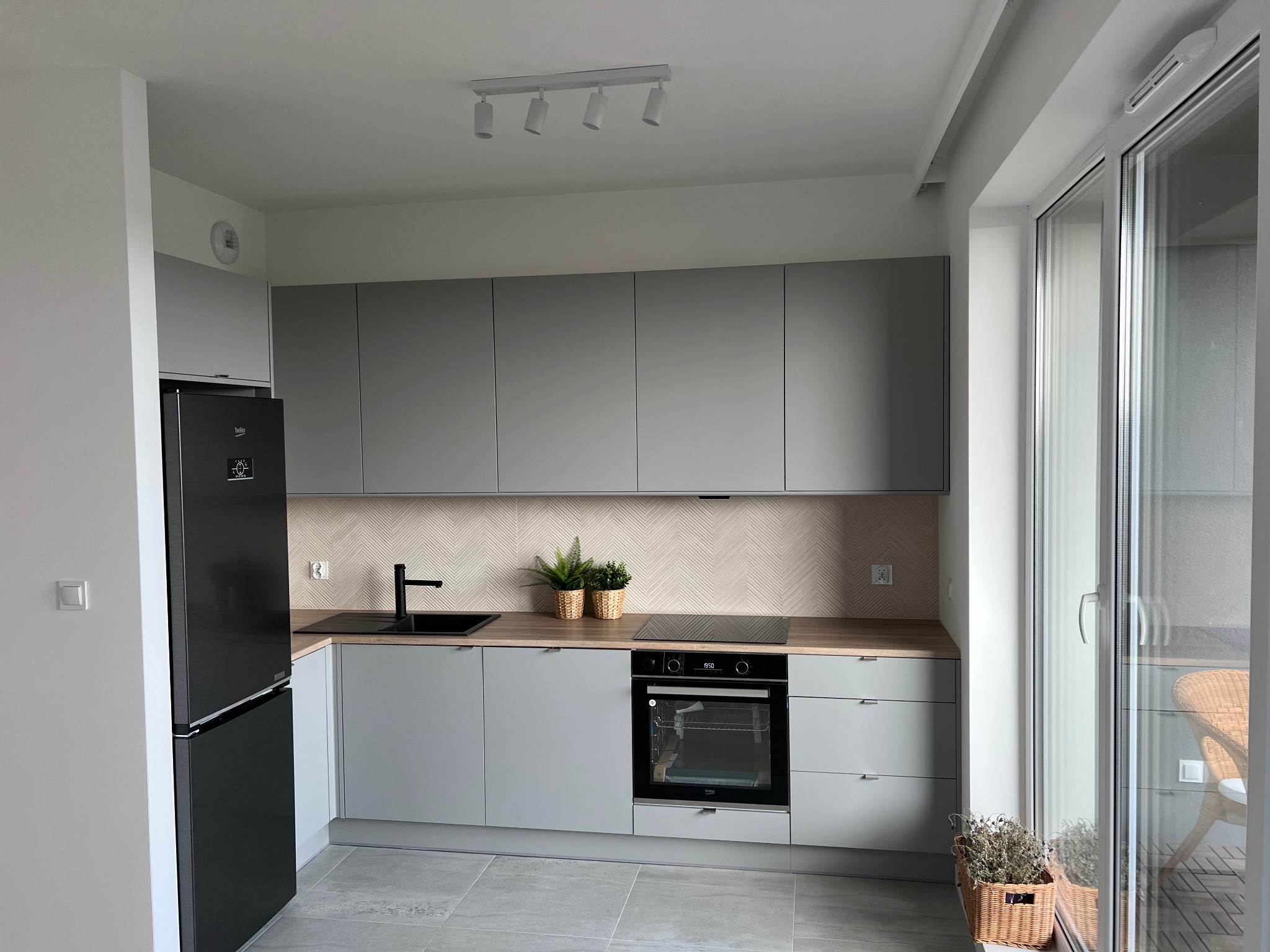 Jasna kuchnia w szarościach: zabudowa meblowa, czarny zlew i sprzęt AGD, płytki w jodełkę nad blatem. Widok na balkon przez okno.