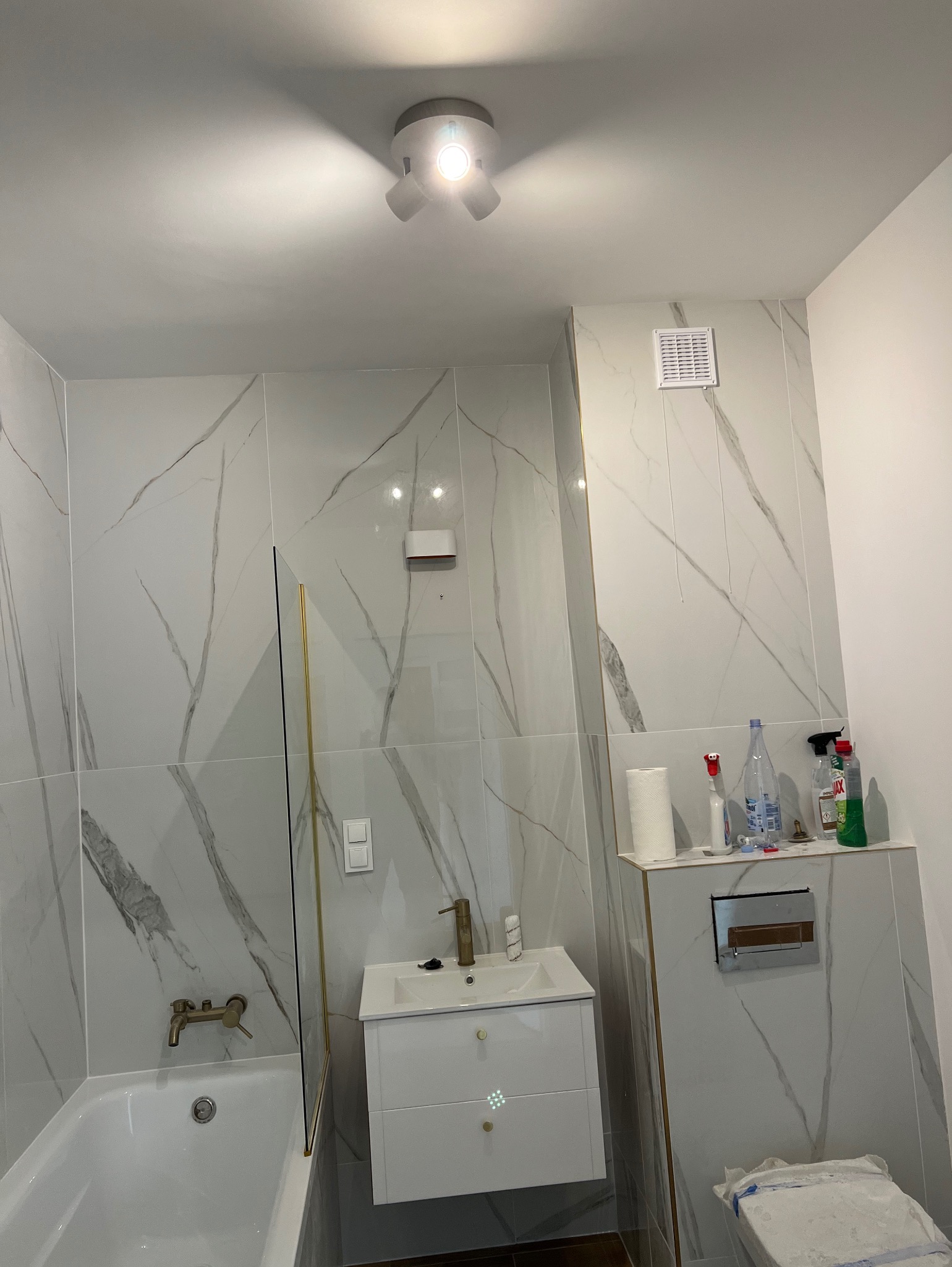 Wykończona łazienka w bieli z marmurowym wzorem płytek, złote akcenty na armaturze i kabinie prysznicowej, nowoczesne oświetlenie.