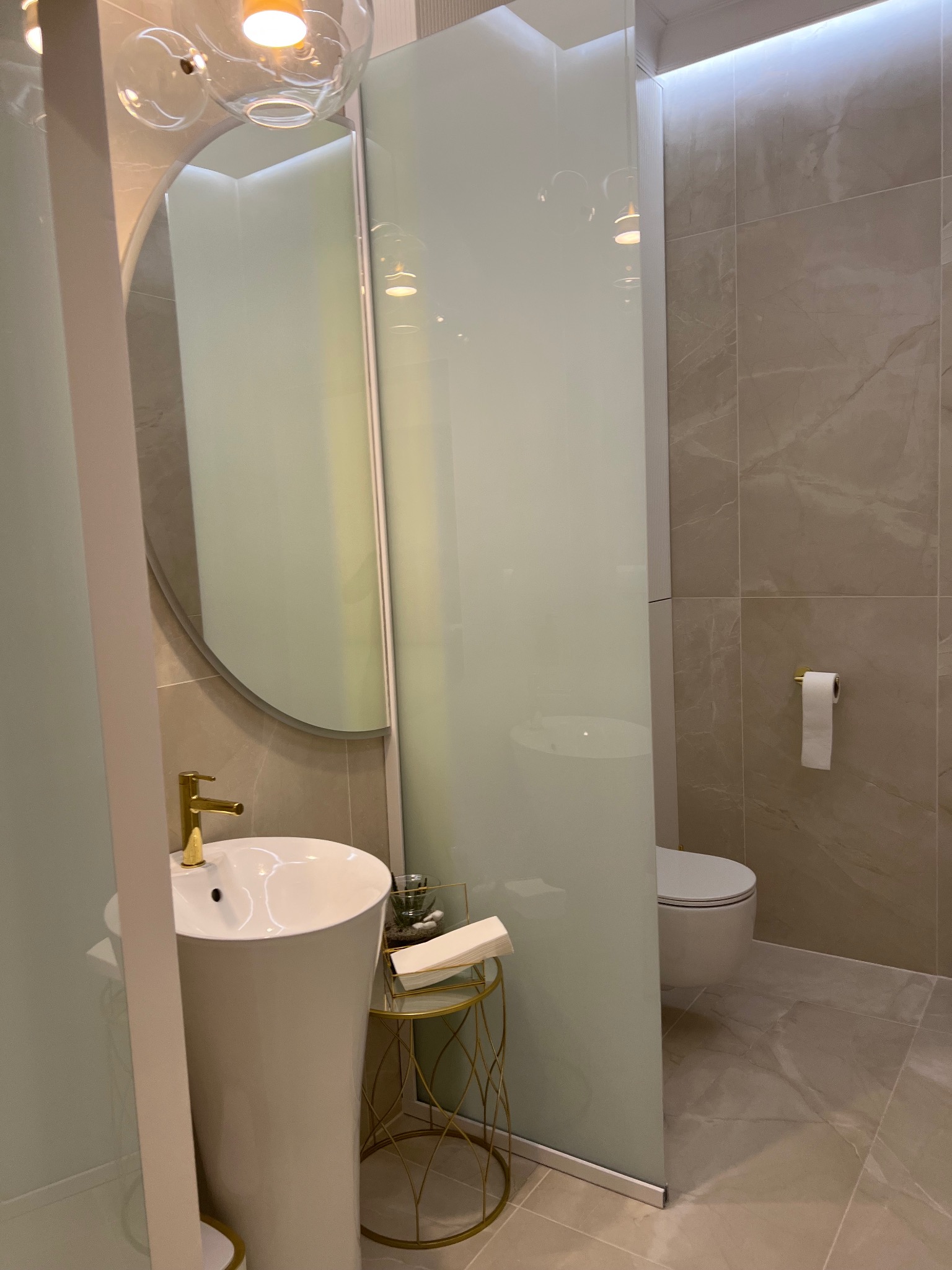 Nowoczesna łazienka z umywalką na postumencie, lustrem i kabiną prysznicową ze szkła. Beżowe płytki imitujące marmur. Minimalistyczny design i złote akcenty.