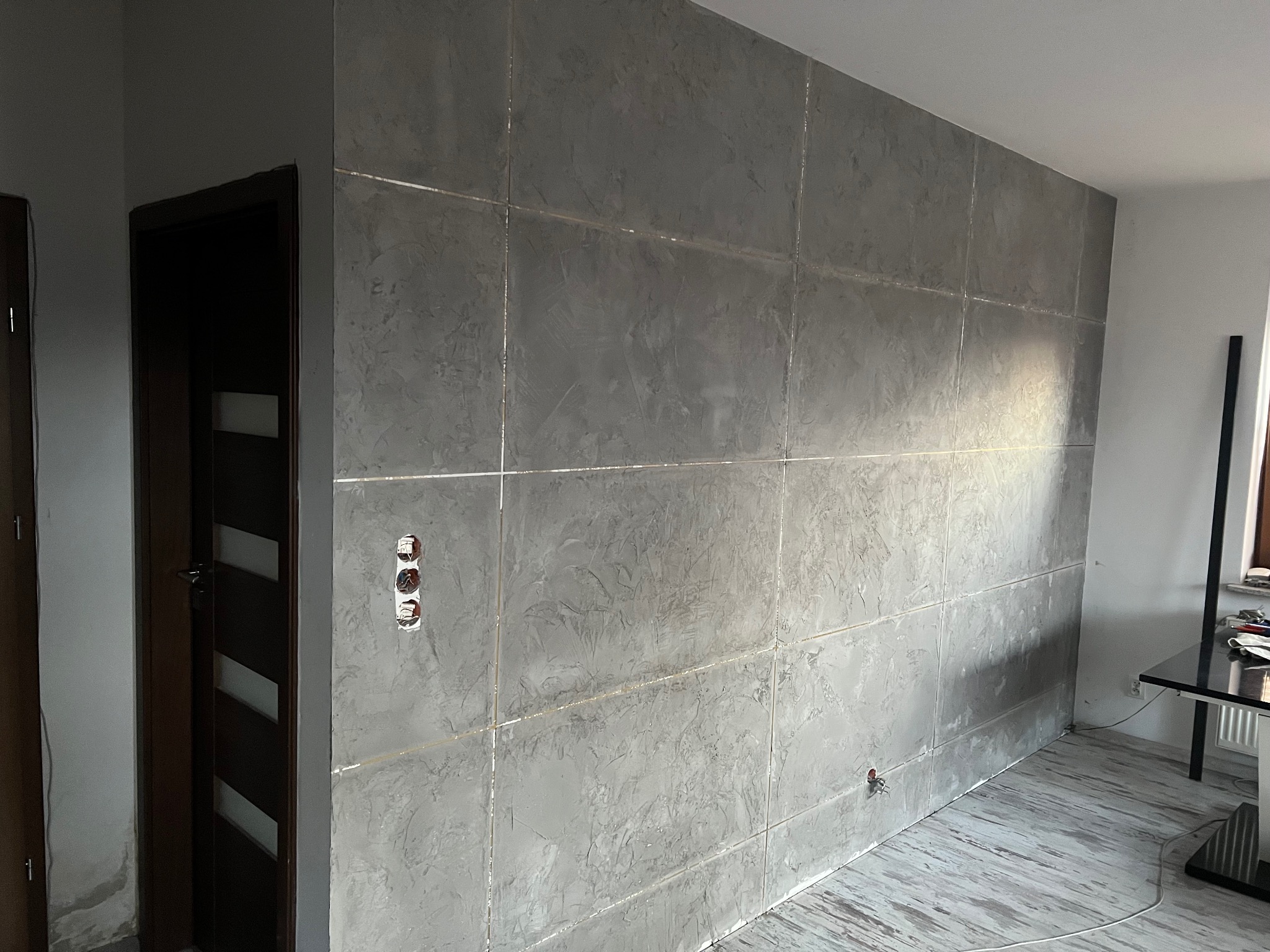 Ściana wykończona dekoracyjną szarą strukturą w panelach, widoczne puszki elektryczne i fragment podłogi z imitacją drewna. W tle drzwi i fragment okna.