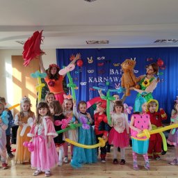 Frajdowersum - Grupa dzieci w kostiumach karnawałowych, z balonami modelowanymi w kształcie zwierząt i kwiatów, pozuje na tle dekoracji z napisem 'Bal Karnawałowy'.