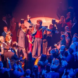 Konferansjer w błyszczącej marynarce i kraciastej spódnicy na scenie, otoczony publicznością robiącą zdjęcia telefonami w czerwono-niebieskim oświetleniu.