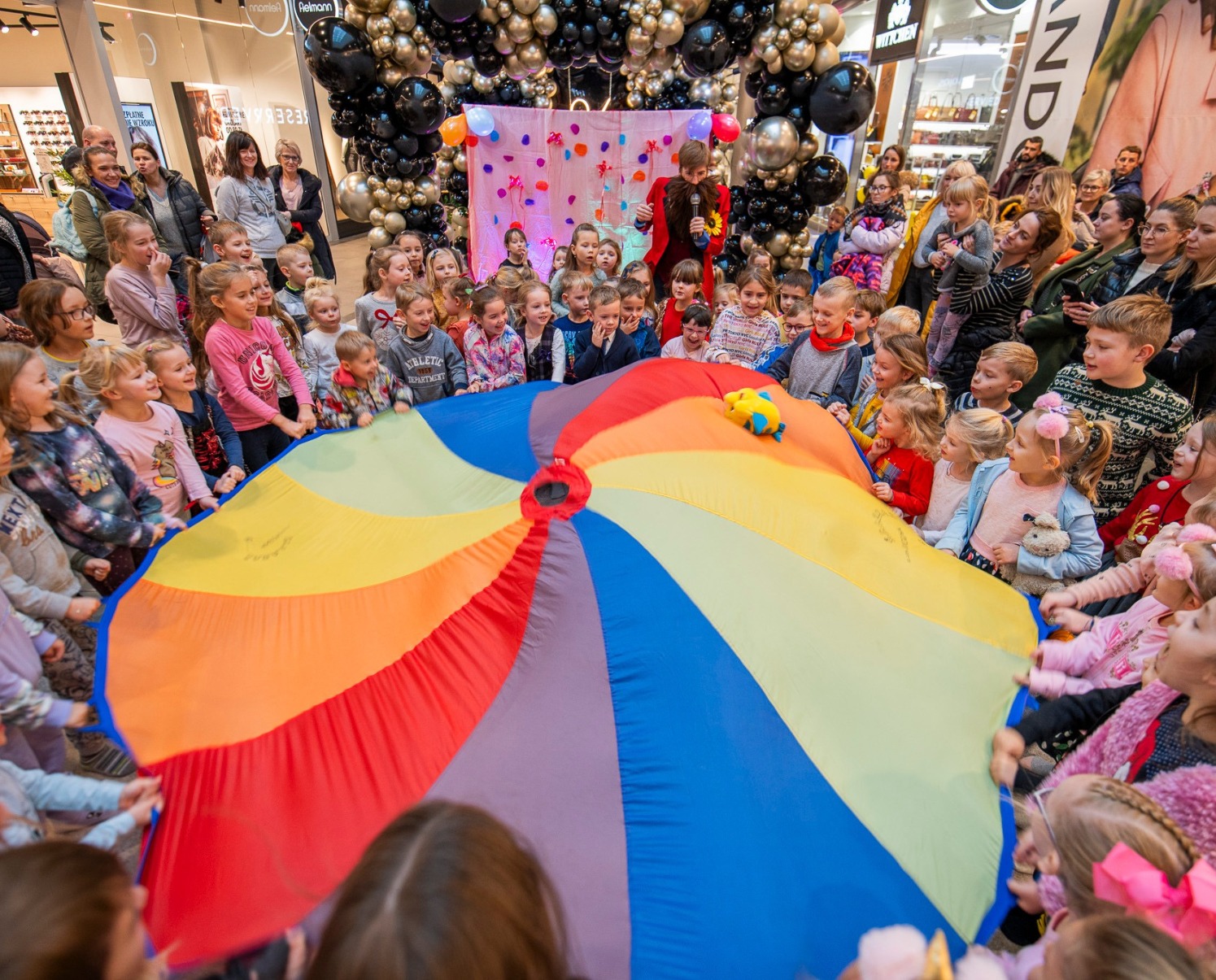 Animator w czerwonym fraku prowadzi zabawy z dziećmi przy kolorowej chuście animacyjnej podczas imprezy w centrum handlowym, tło z balonów.