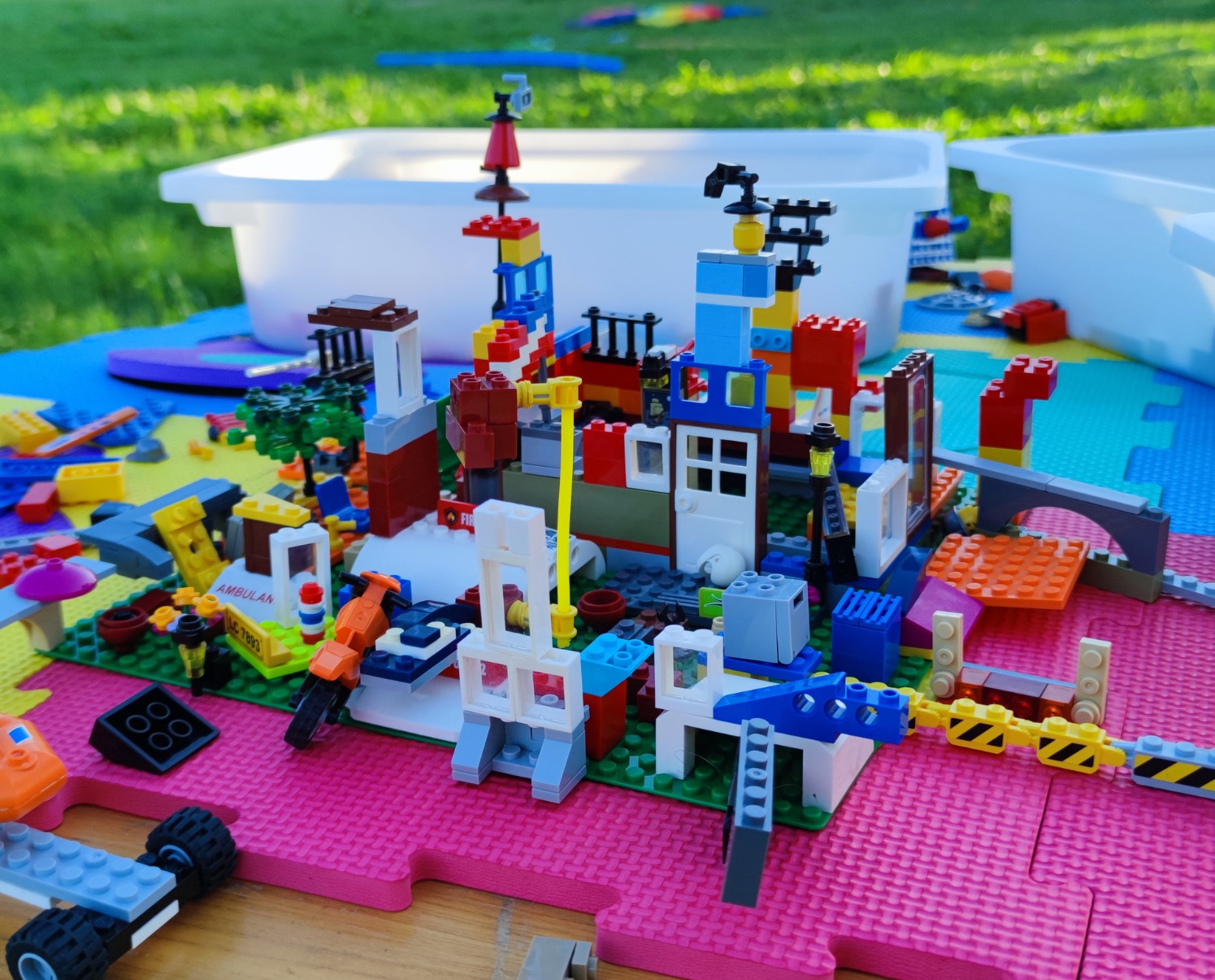 Kolorowa konstrukcja z klocków LEGO na macie piankowej, z elementami miasta, pojazdami i budynkami. W tle białe pojemniki i trawa.