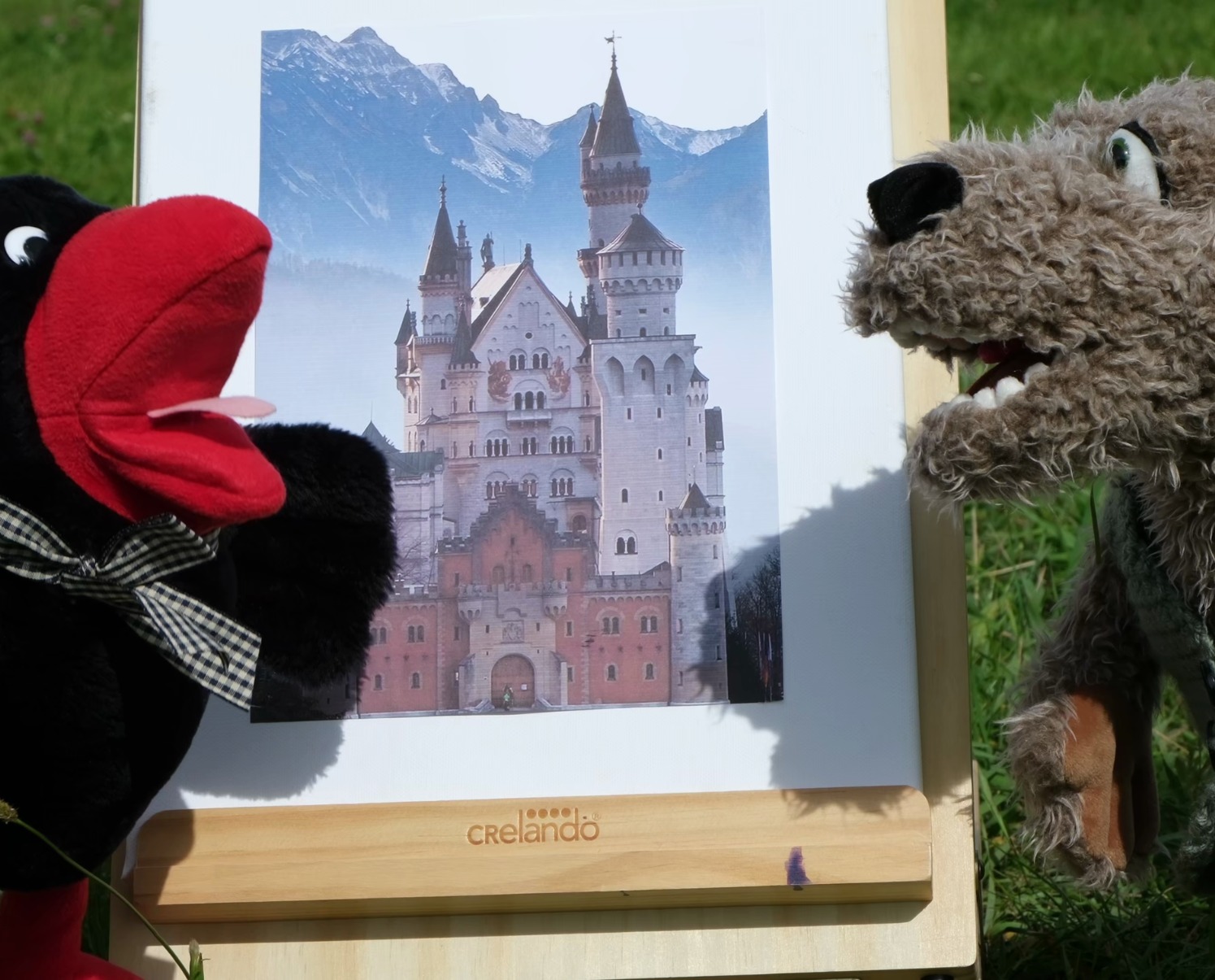 Teatrzyk kukiełkowy z zamkiem Neuschwanstein w tle, na zielonej trawie. Kukiełki ptaka i wilka, na drewnianej sztaludze firmy Crelando. Scena z warsztatów teatralnych dla dzieci.
