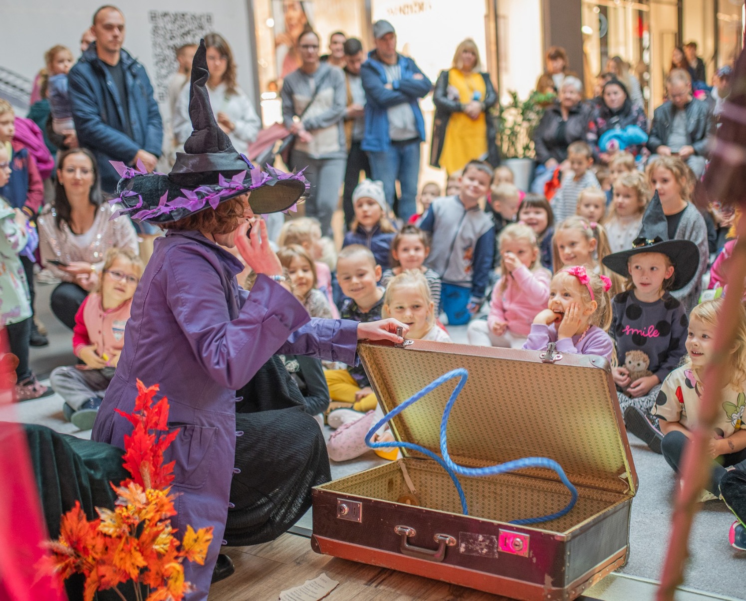 Animator w stroju wiedźmy prezentuje rekwizyty z walizki grupie dzieci siedzących na podłodze, z dorosłymi obserwatorami w tle.