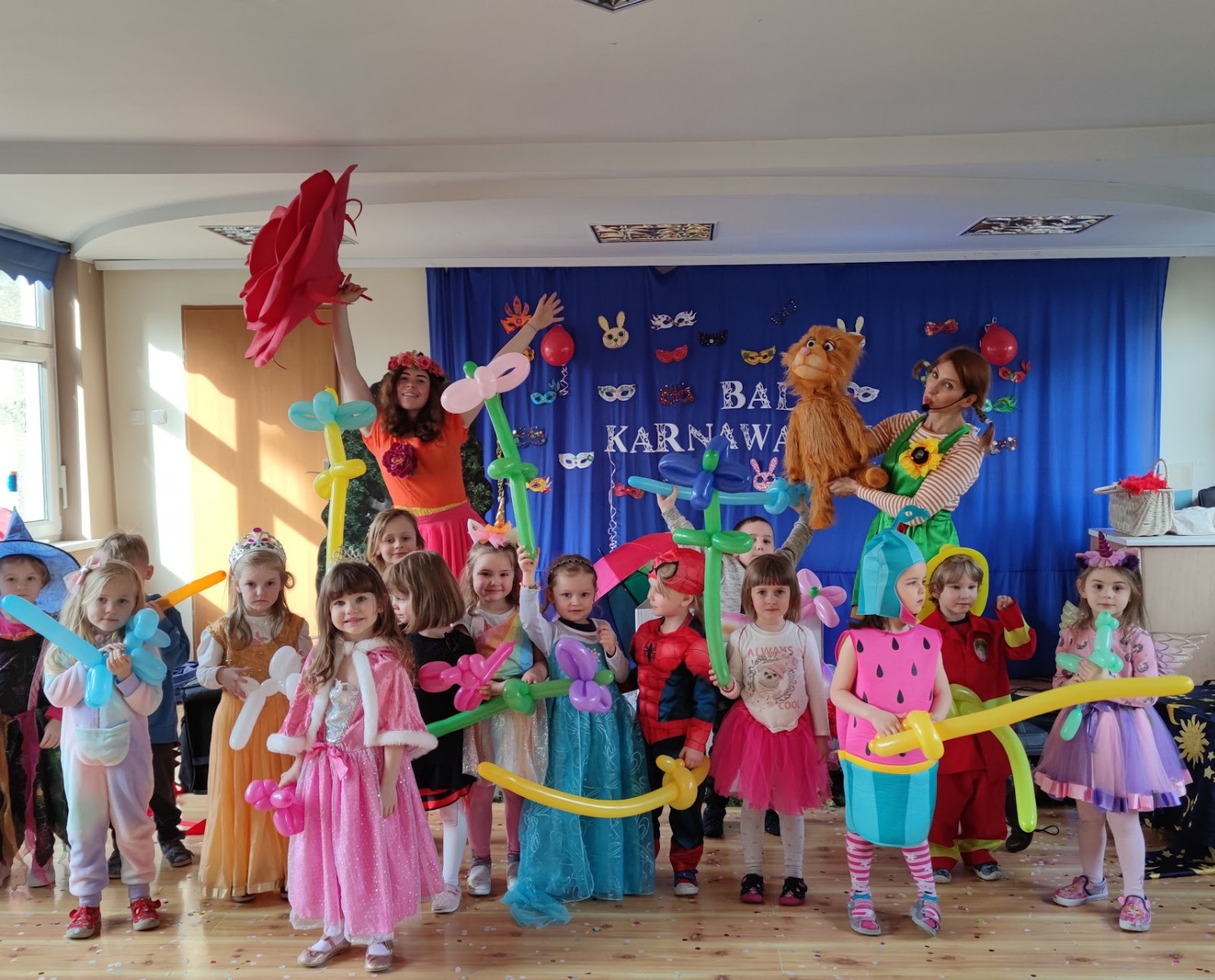 Grupa dzieci w kostiumach karnawałowych, z balonami modelowanymi w kształcie zwierząt i kwiatów, pozuje na tle dekoracji z napisem 'Bal Karnawałowy'.