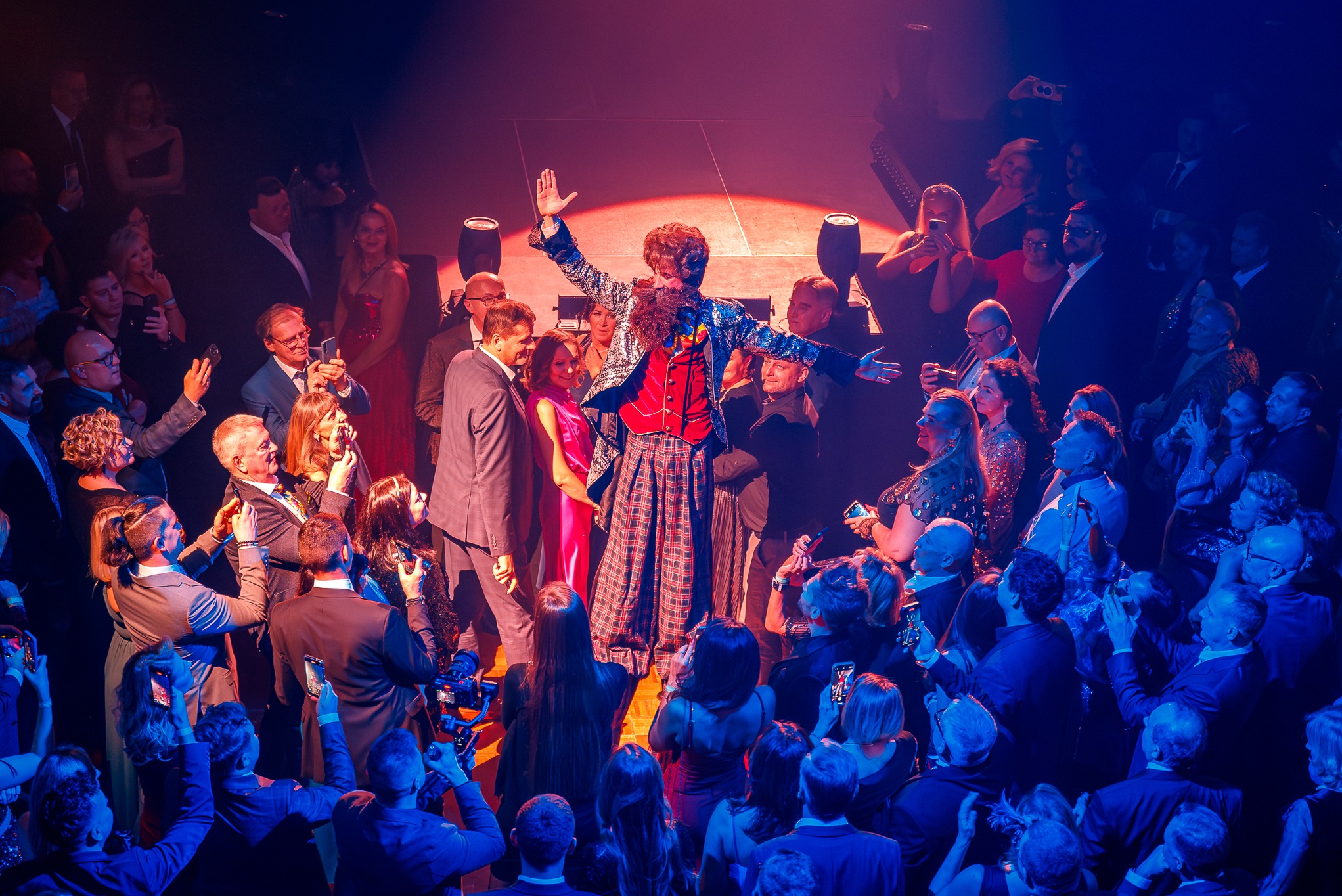 Konferansjer w błyszczącej marynarce i kraciastej spódnicy na scenie, otoczony publicznością robiącą zdjęcia telefonami w czerwono-niebieskim oświetleniu.