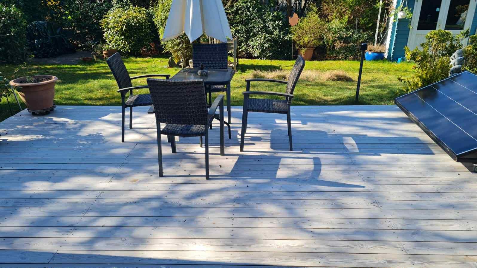 Bielona deska tarasowa z ciemnymi meblami ogrodowymi i panelem słonecznym w tle zielonego ogrodu, oświetlona promieniami słońca.