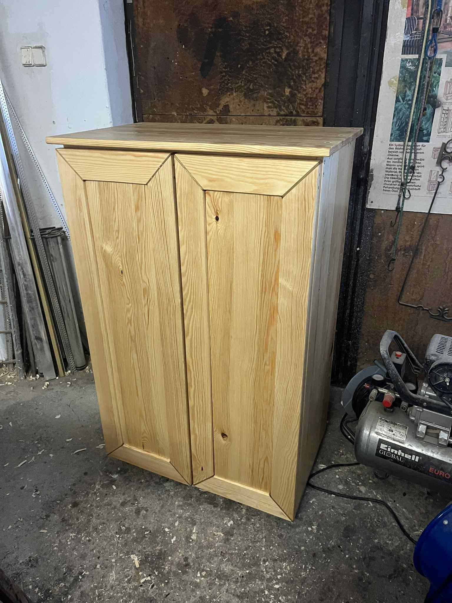 Drewniana szafka dwudrzwiowa z surowego drewna sosnowego, z widocznymi słojami, stoi na betonowej podłodze w warsztacie, obok kompresora.