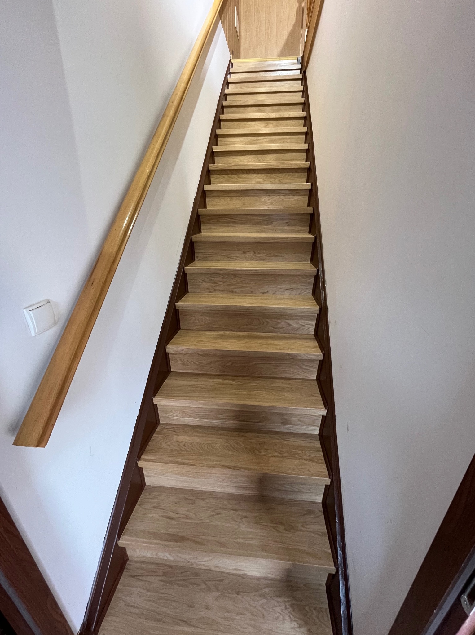 Jasne, drewniane schody z lakierowaną poręczą, widziane z dołu, w białym korytarzu. Ciemnobrązowe wykończenie stopni. Widoczny włącznik światła na ścianie.
