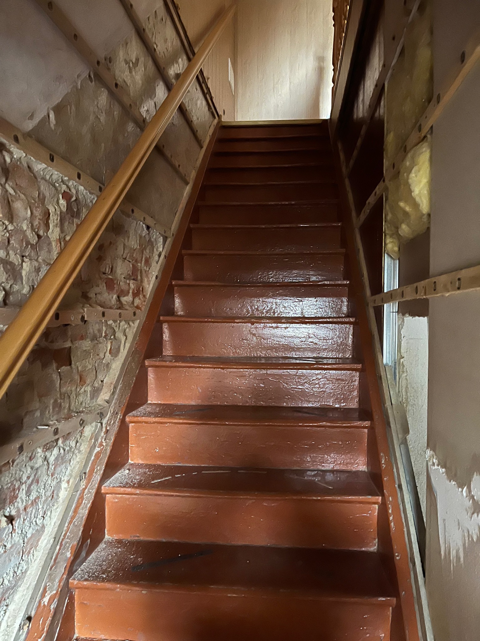Schody drewniane w trakcie renowacji, widoczne ślady farby i odsłonięta cegła na ścianie. Ujęcie z dołu, podkreślające etap prac wykończeniowych.