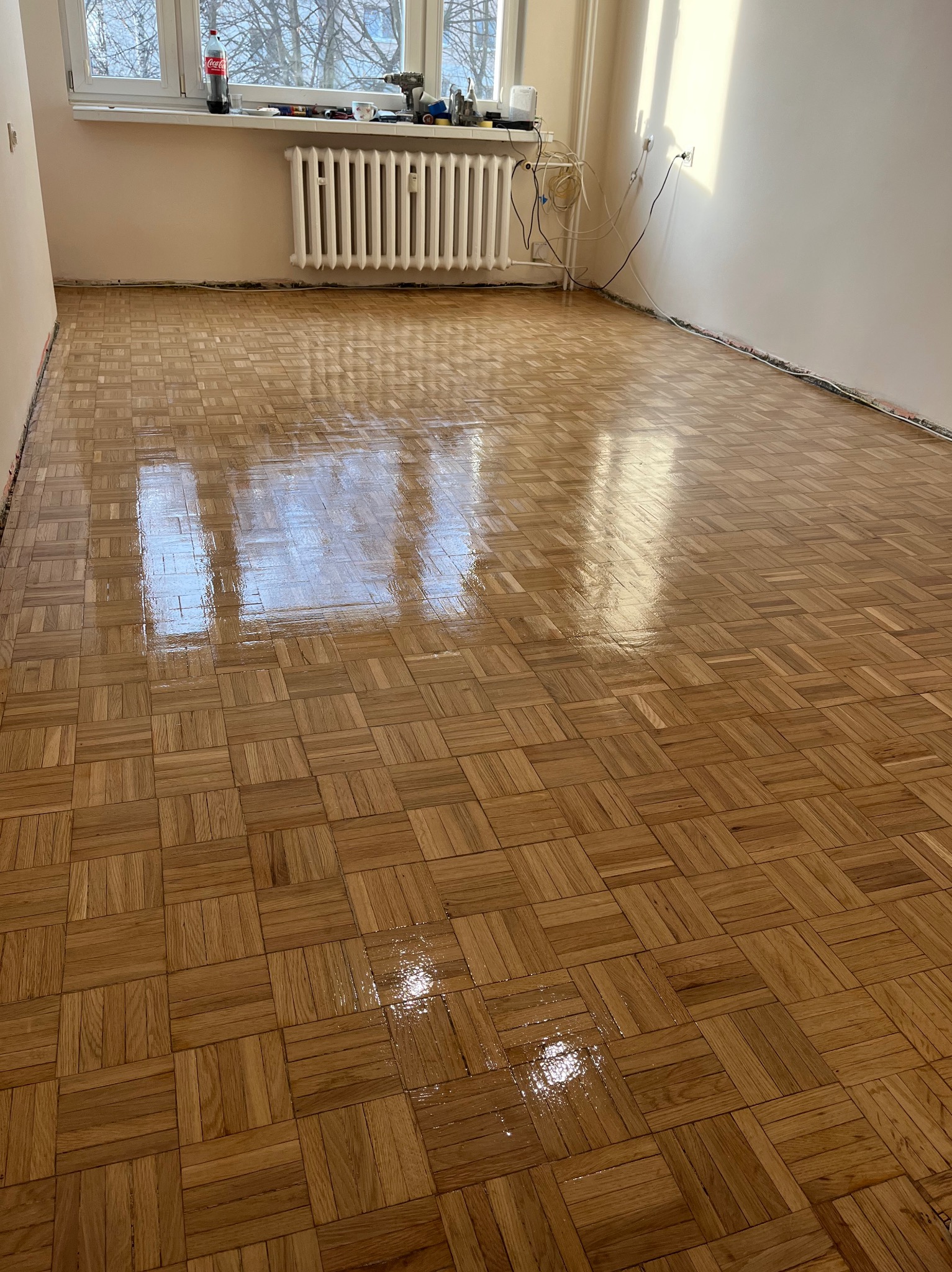 Błyszcząca, odnowiona podłoga parkietowa w jodełkę, po cyklinowaniu i lakierowaniu. Widoczne refleksy światła, jasne ściany i okno w tle. Brak listew przypodłogowych.