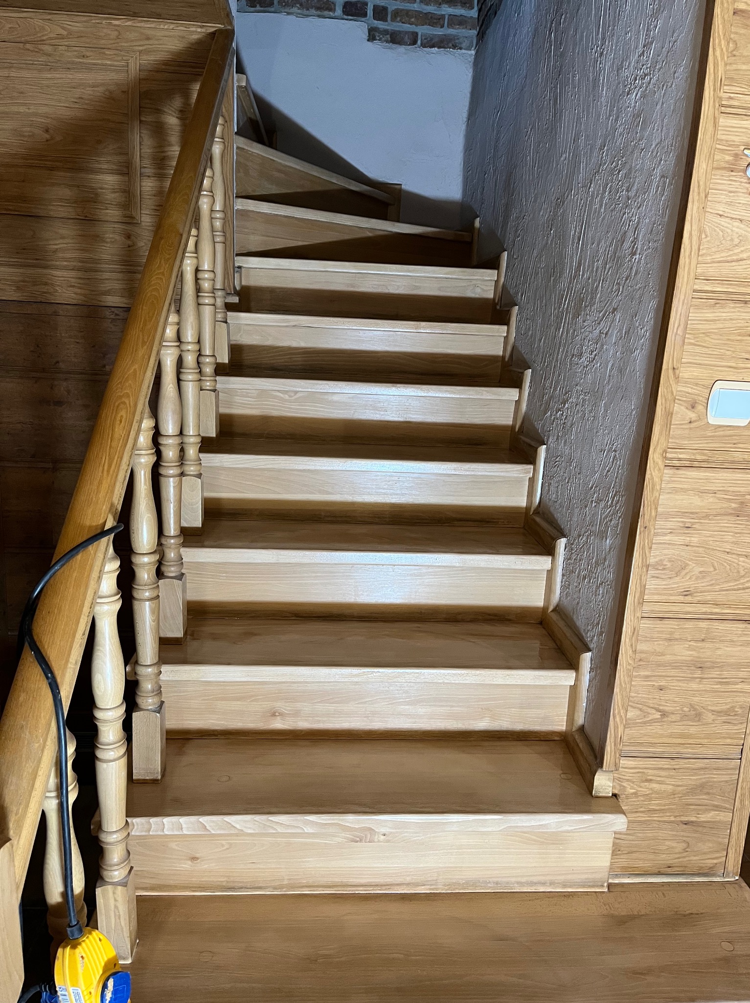 Jasne, drewniane schody z balustradą, widok z dołu. Naturalne oświetlenie podkreśla strukturę drewna. Ściana z fakturą i fragment cegły w tle. Widoczny kabel z żółtą wtyczką.