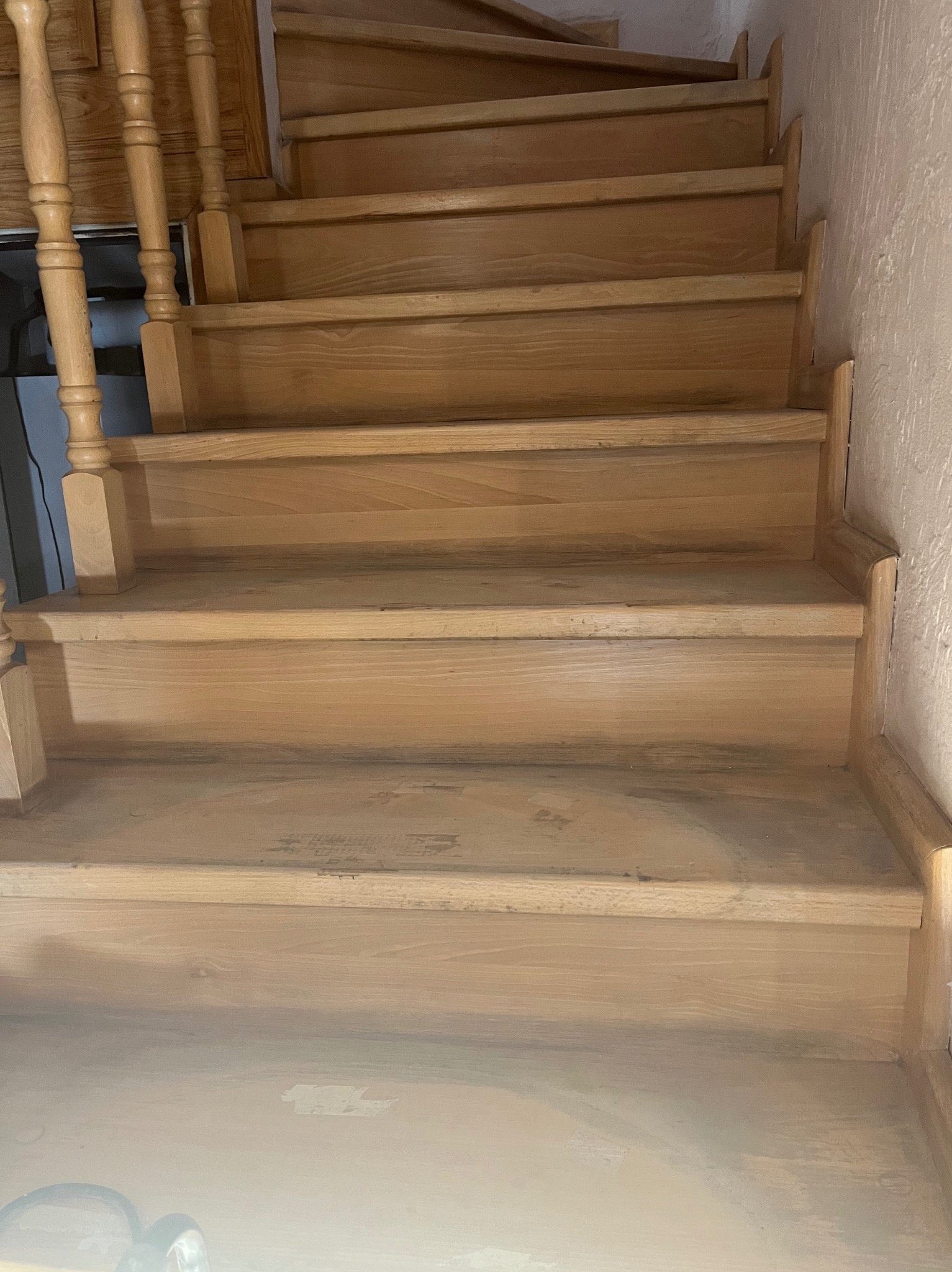 Jasne, drewniane schody z balustradą, widziane z dołu. Widoczne ślady użytkowania na stopniach, sugerujące potrzebę renowacji. Naturalne oświetlenie podkreśla strukturę drewna.