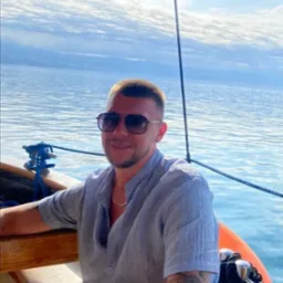 Uśmiechnięty mężczyzna w okularach przeciwsłonecznych, siedzący na łodzi na tle morza i błękitnego nieba z chmurami. Widoczny tatuaż na ramieniu.